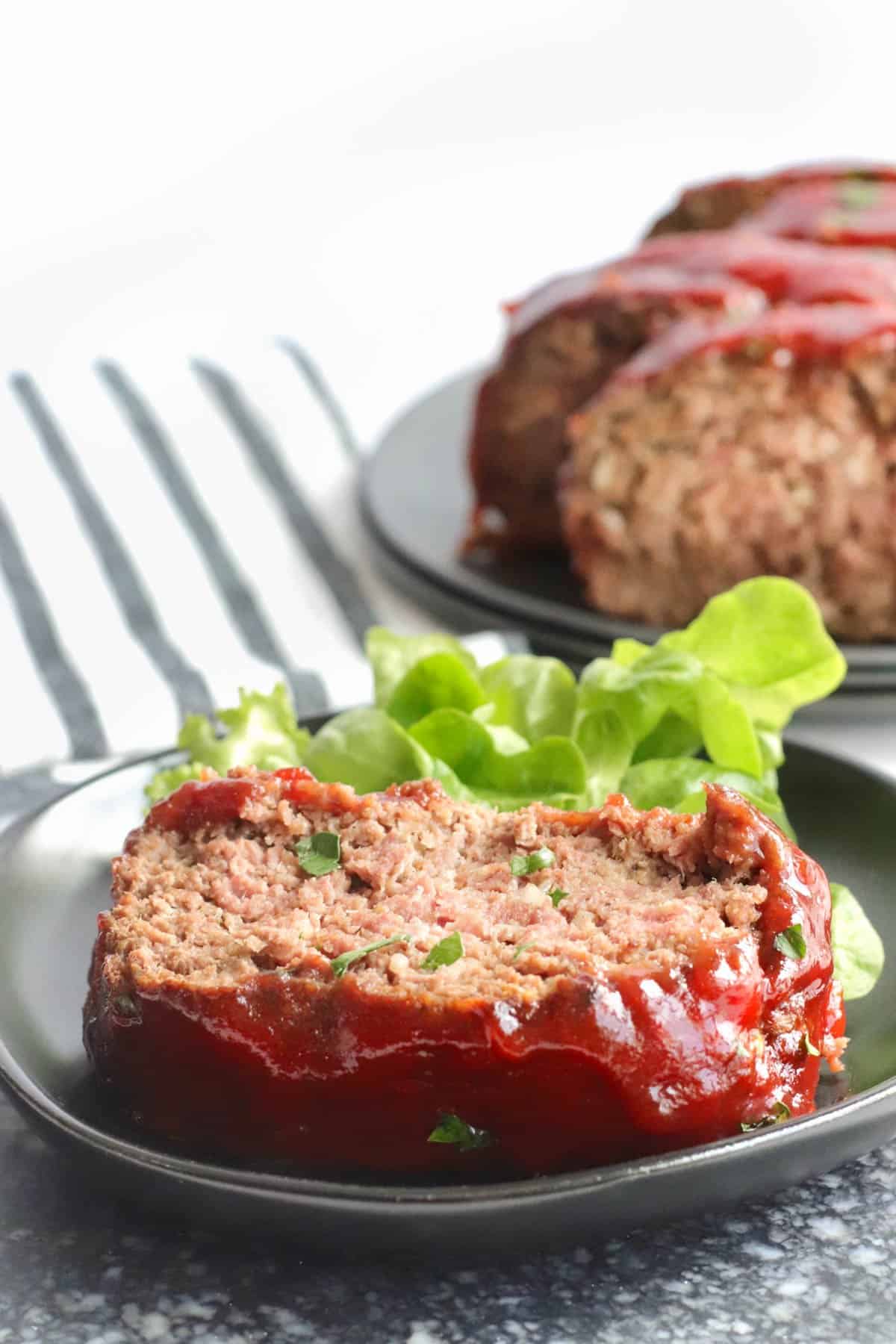 A piece of meatloaf laying flat on a black plat with a leaf of green lettuce.