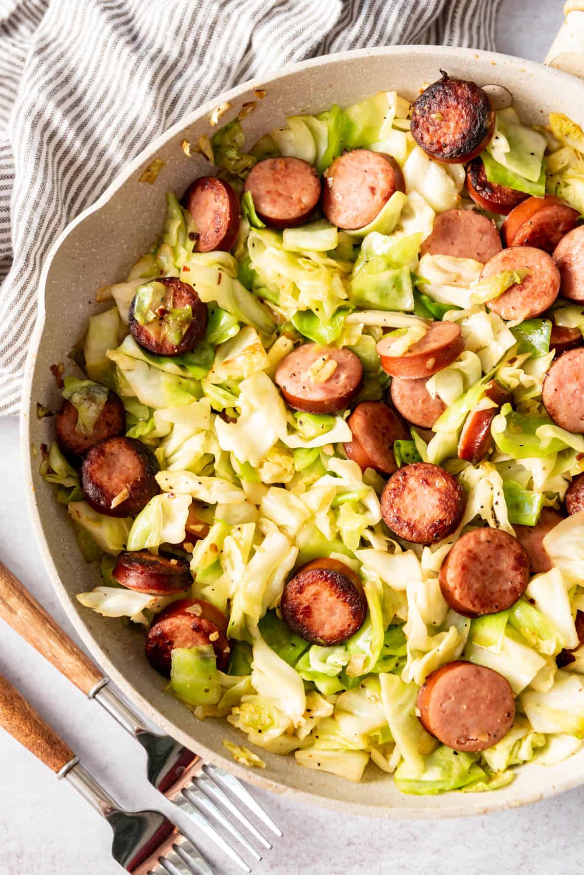 Sauteed cabbage and kielbasa sausage in a pan next to forks and a linen napkin.