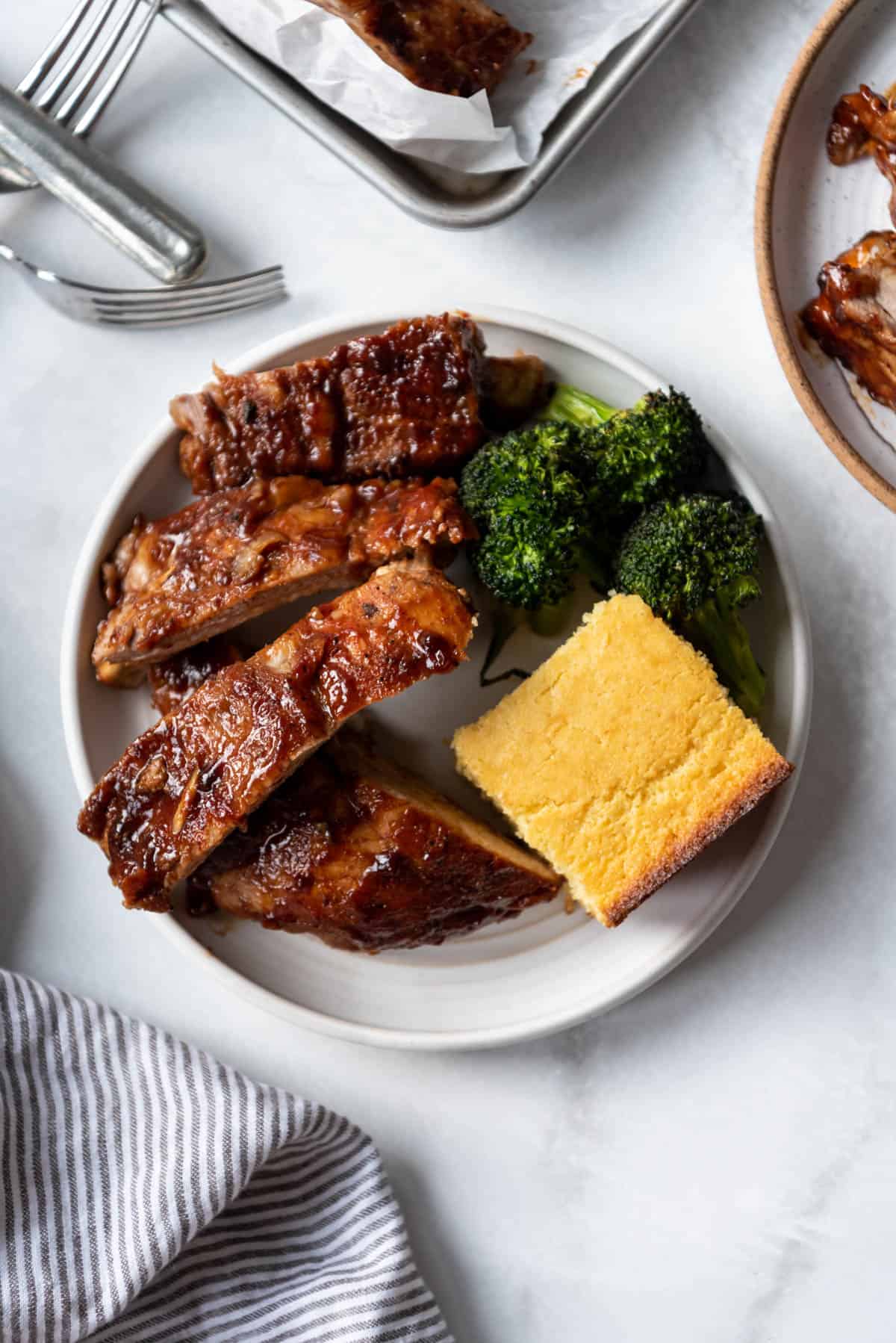 A white plate with four baby back ribs, cornbread, and roasted broccoli.