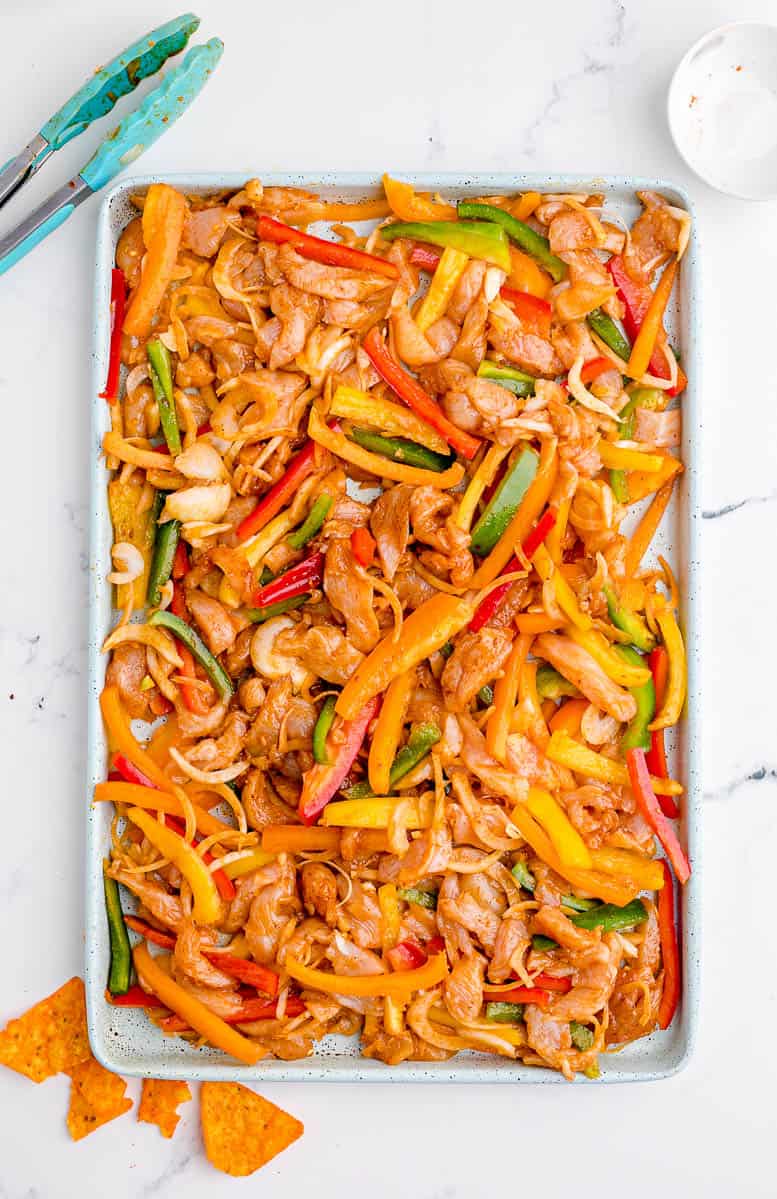 Uncooked sliced chicken, bell peppers, and onions on a baking sheet.