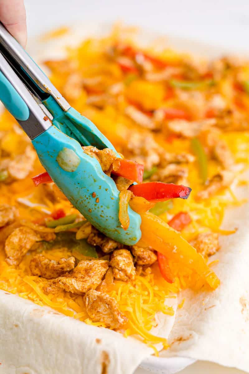 Adding cooked fajita veggies and chicken to grated cheese on flour tortillas for oven quesdillas.