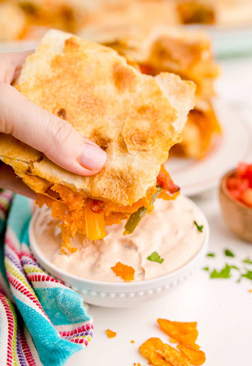 A hand dipping a baked chicken quesadilla into a creamy dipping sauce.