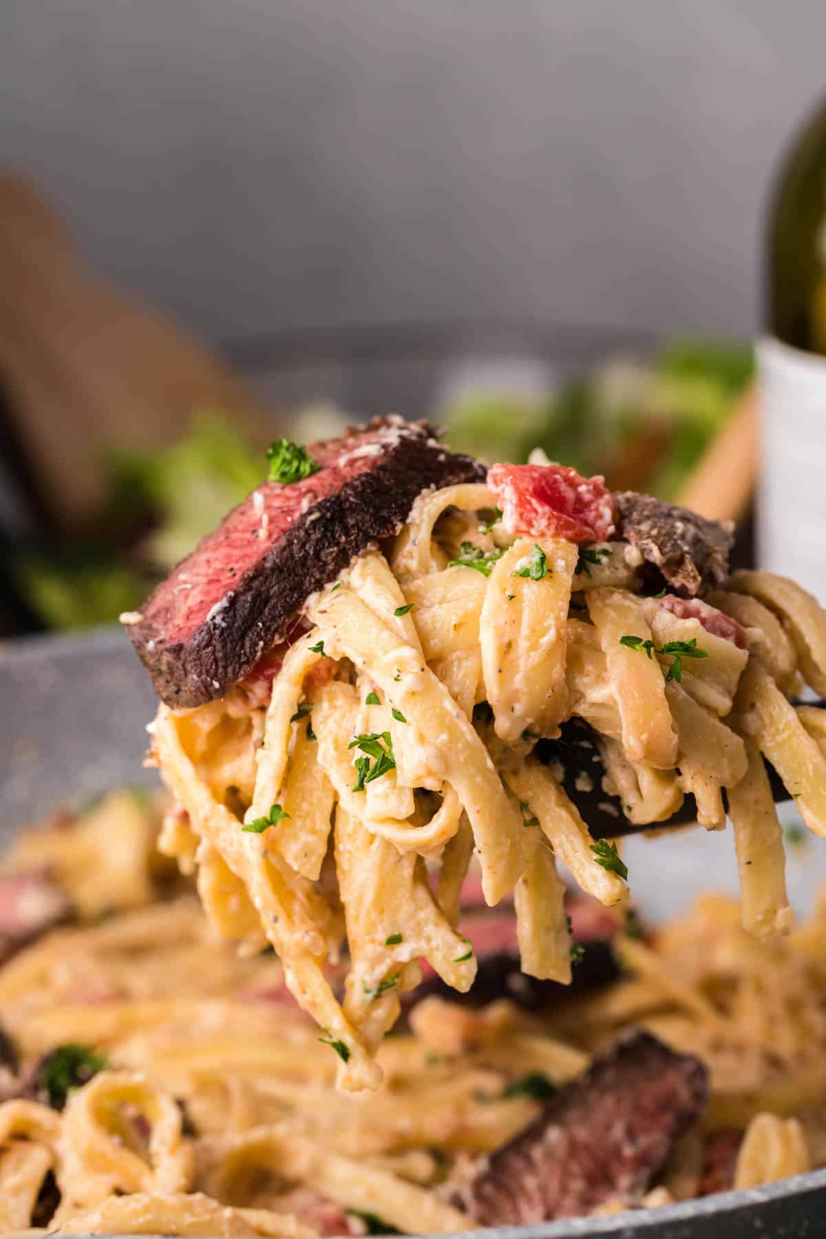 A spoonful of fettuccine alfredo noodles and sliced steak being lifted out of the pan.