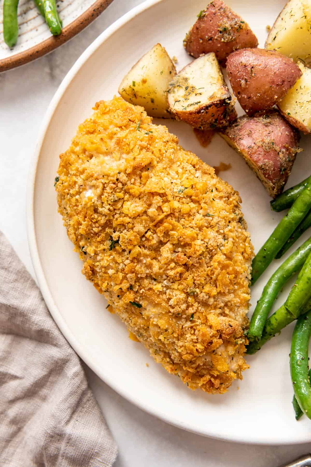 A cornflake and ranch seasoning crusted chicken breast on a plate with vegetables.