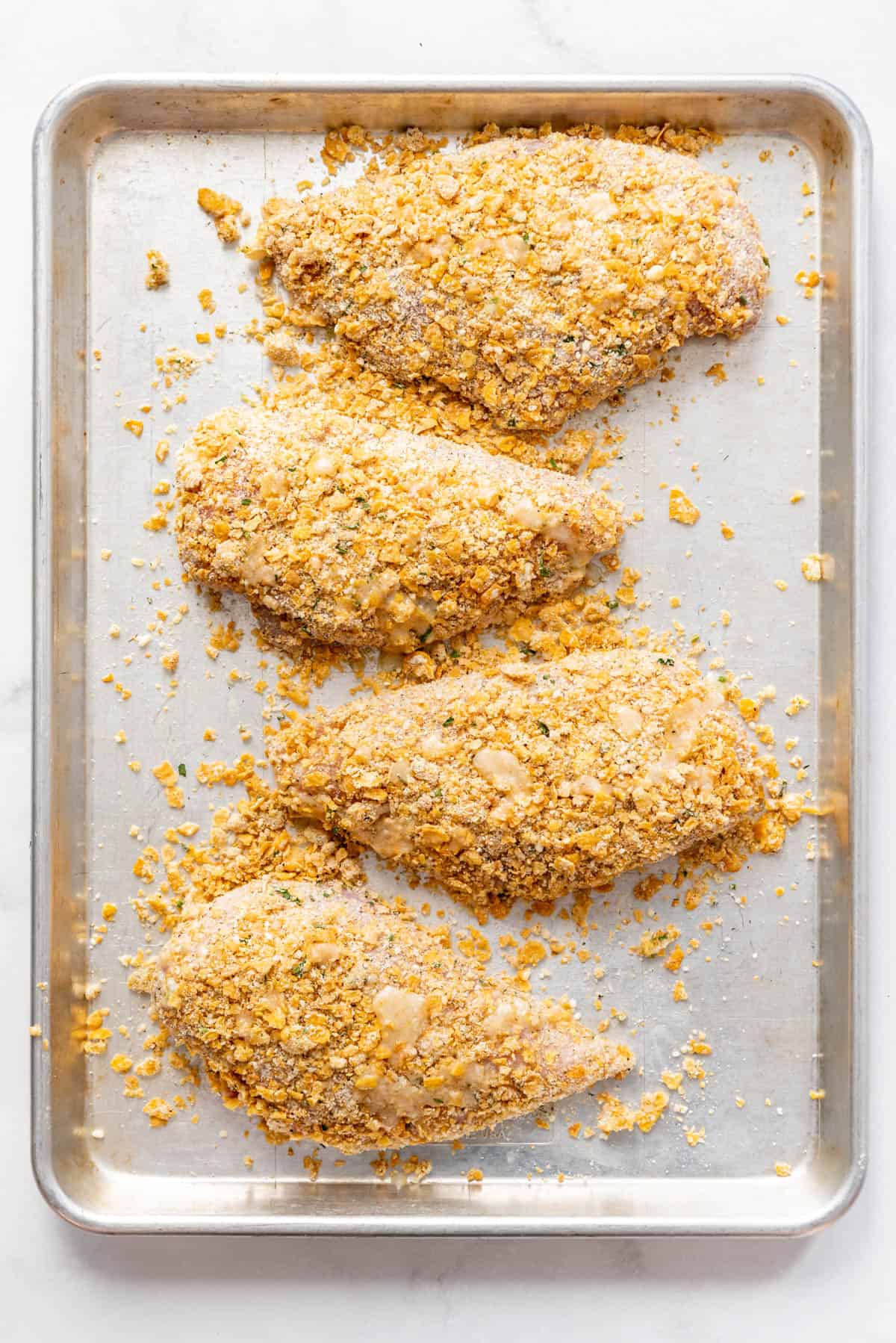 Four breaded chicken breasts on a baking sheet ready to go into the oven.