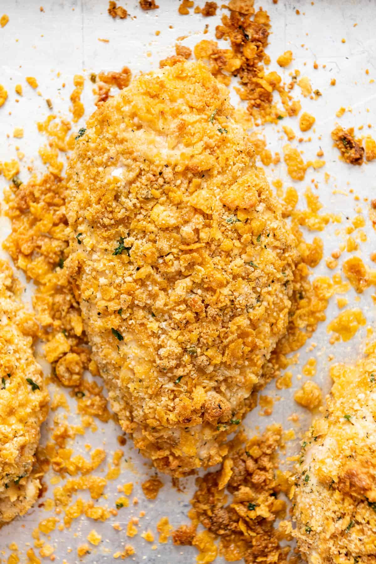 A close image of a piece of baked chicken with ranch seasoning and cornflake breading on a baking sheet.
