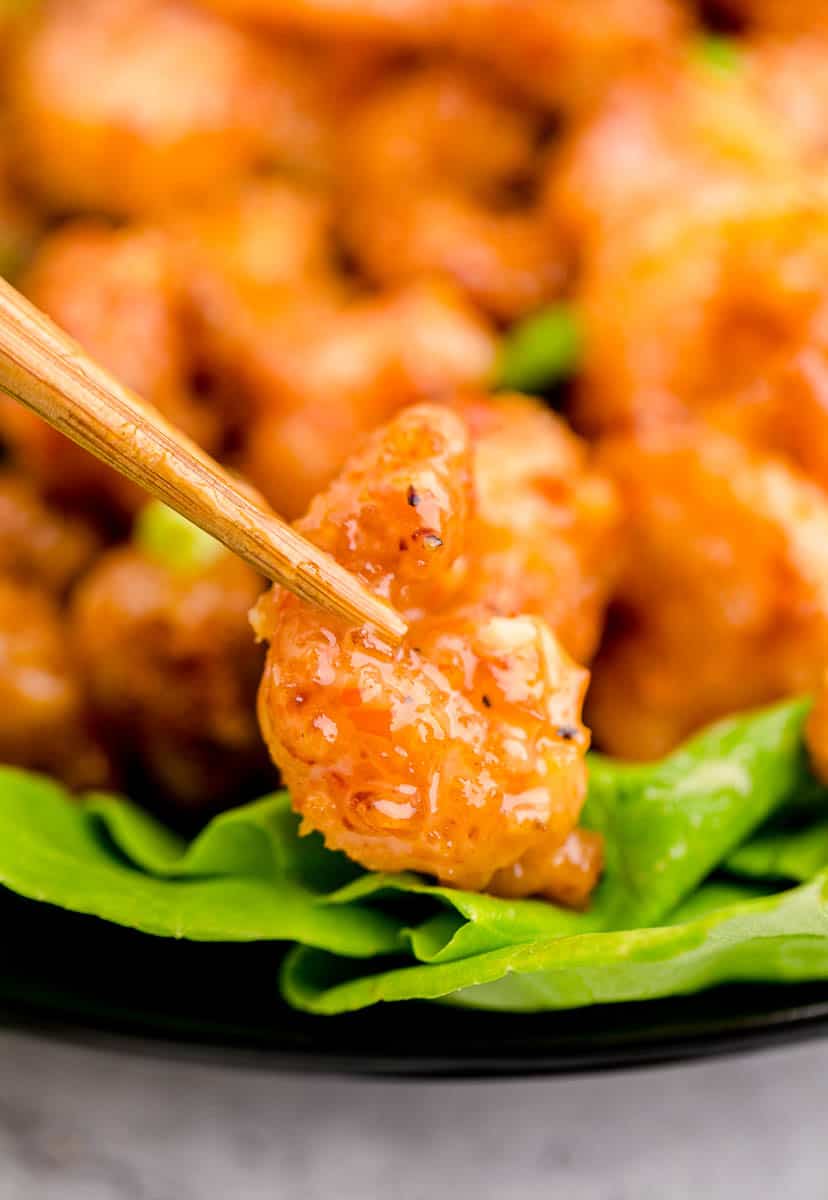 A pair of chopsticks holding a single bang bang shrimp with a bowl full of shrimp in the background