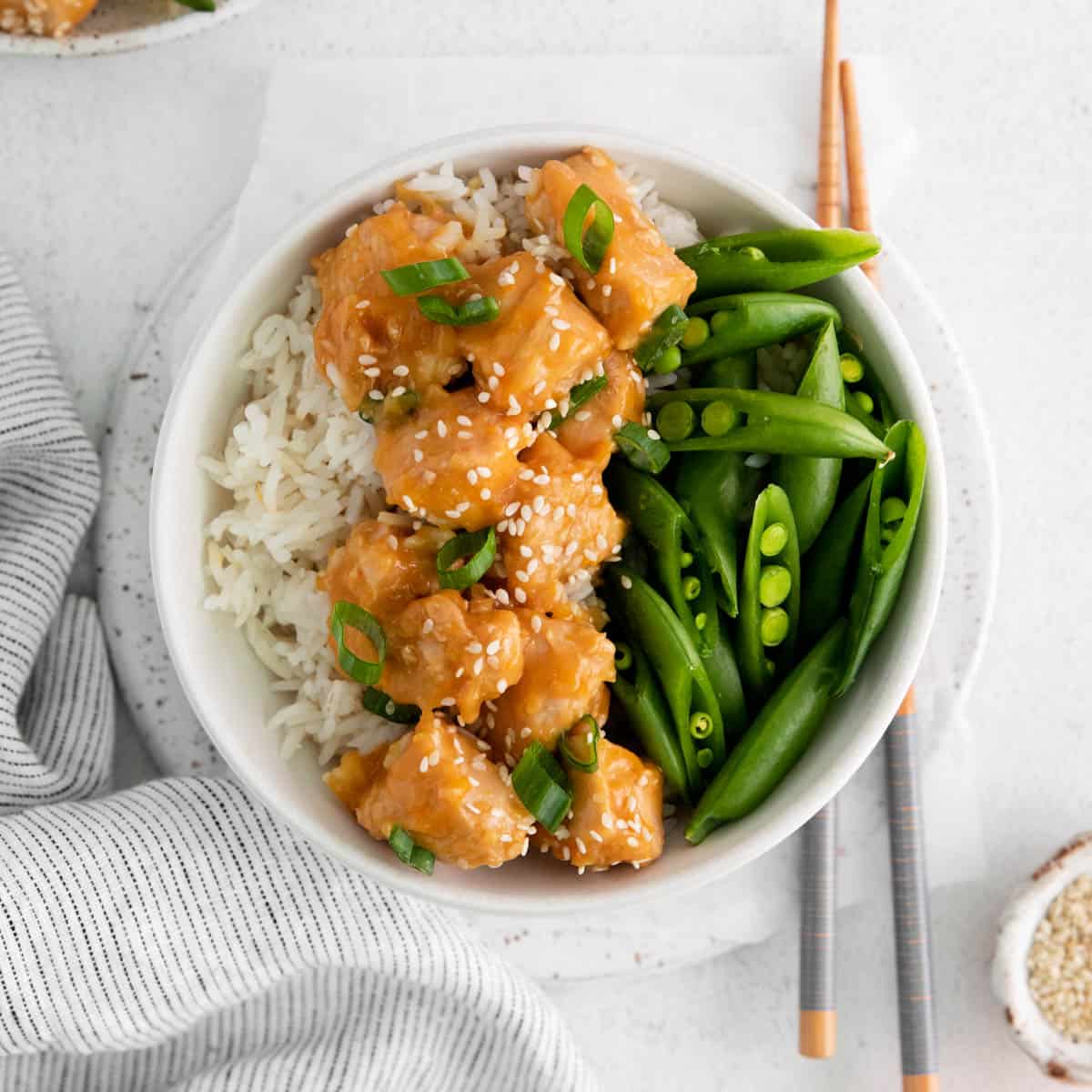 Pieces of orange chicken with sesame seeds and green onions on top in a bowl with rice and veggies..