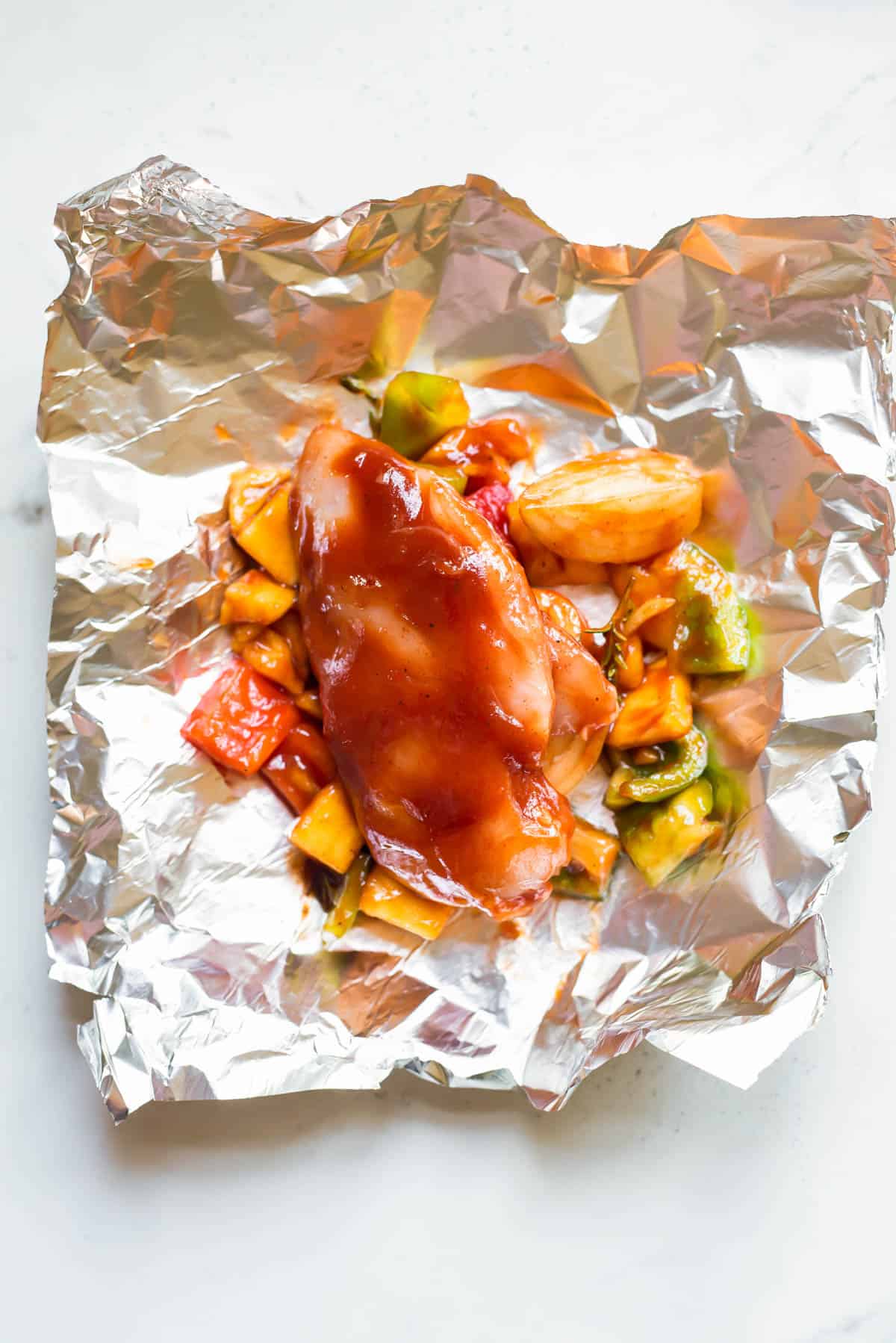 A chicken breast covered in bbq sauce resting on top of chopped pineapple and vegetables ready to be wrapped in aluminum foil.