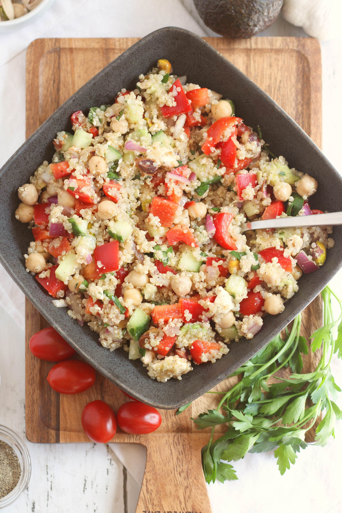 Quinoa salad is displayed in a dark, rectangular bowl with tomatoes and herbs scattered around the bowl. A spoon is visible in the salad. 