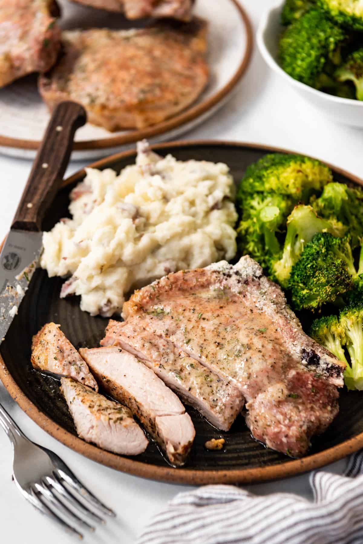 A partially sliced bone-in ranch pork chop on a plate with mashed potatoes and broccoli.