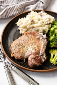 A baked pork chop on a dark plate next to mashed potatoes and roasted broccoli.