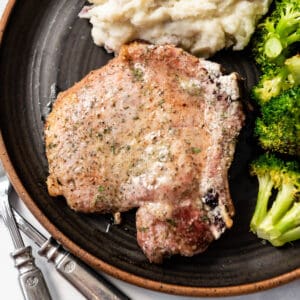 A baked pork chop on a dark plate next to mashed potatoes and roasted broccoli.