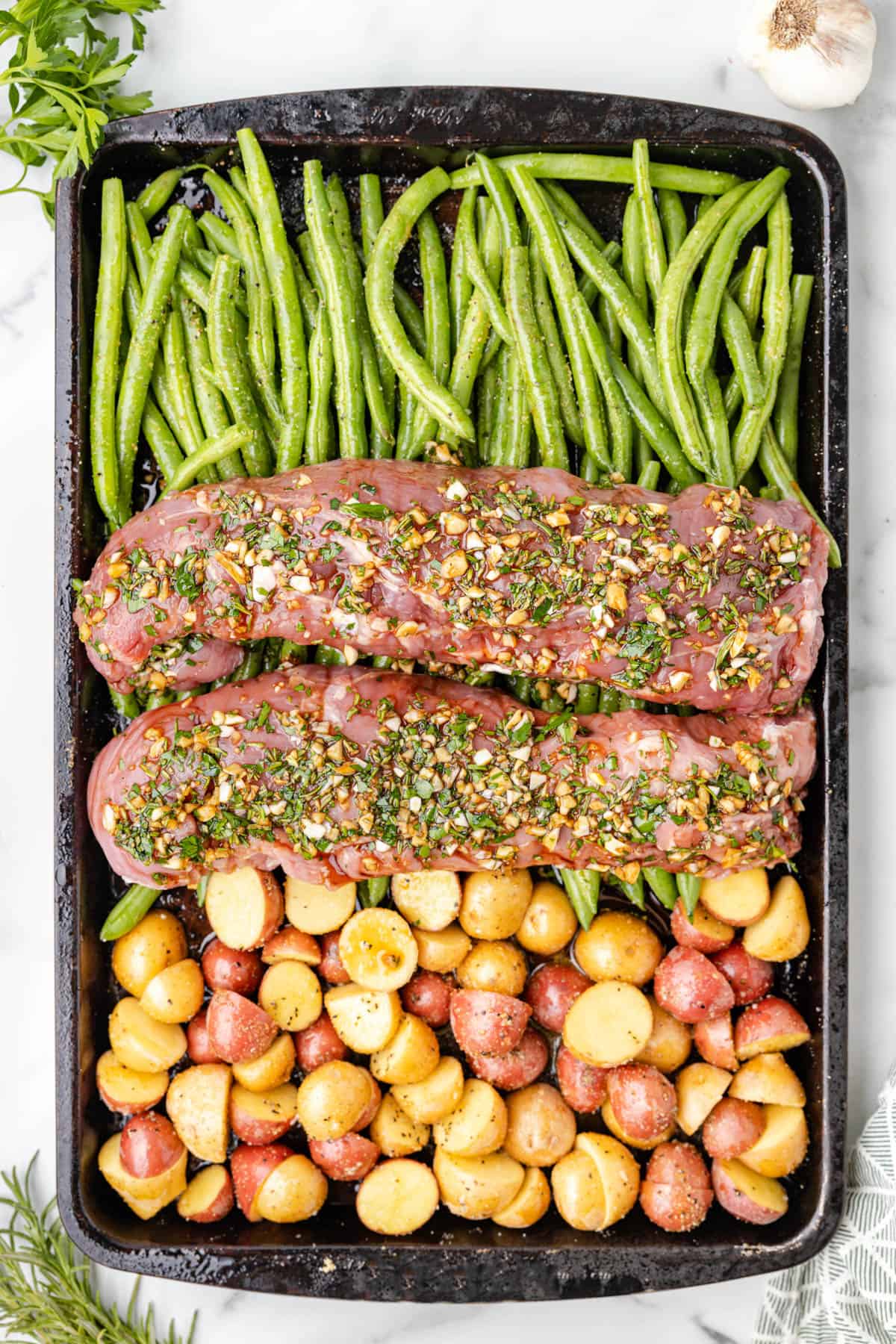 Two pork tenderloin roasts covered in an herb sauce on a sheet pan with potatoes and green beans for roasting.
