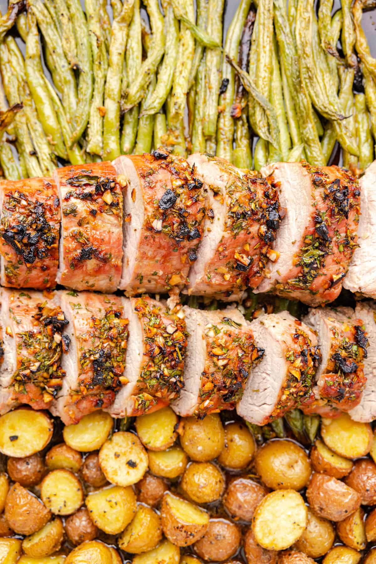 Two sliced pork tenderloin roasts on a bed of roasted potatoes and green beans.