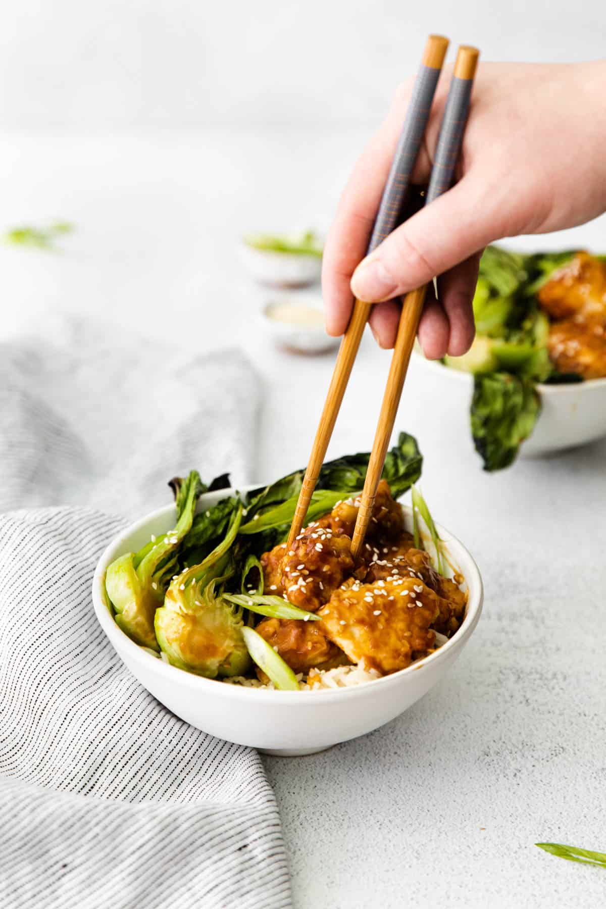 A hand using chopsticks to pick up sticky sesame chicken from a bowl.