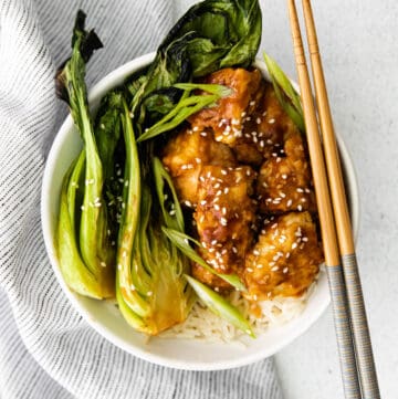 A bowl of sticky sesame chicken, bok choy, and rice with chop sticks.