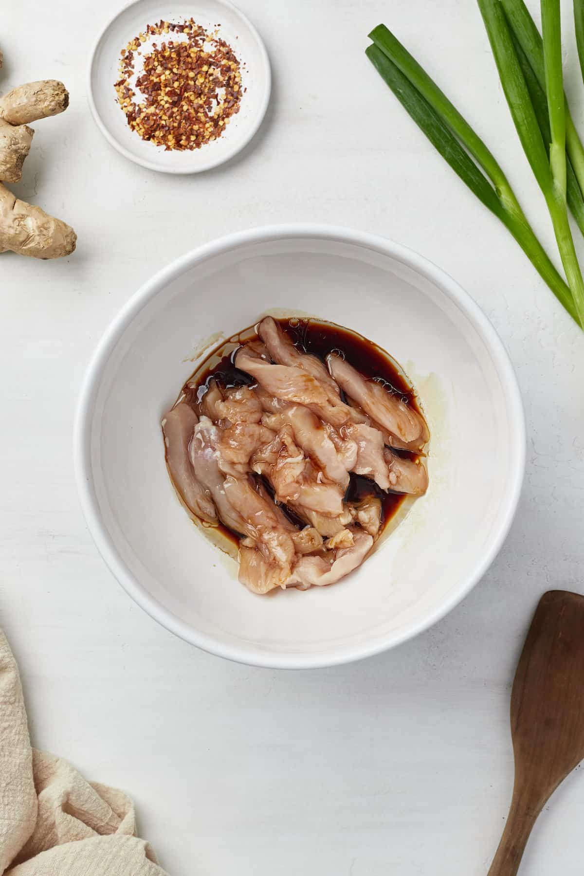 Thin strips of raw chicken breast in a bowl with soy sauce and sesame oil.