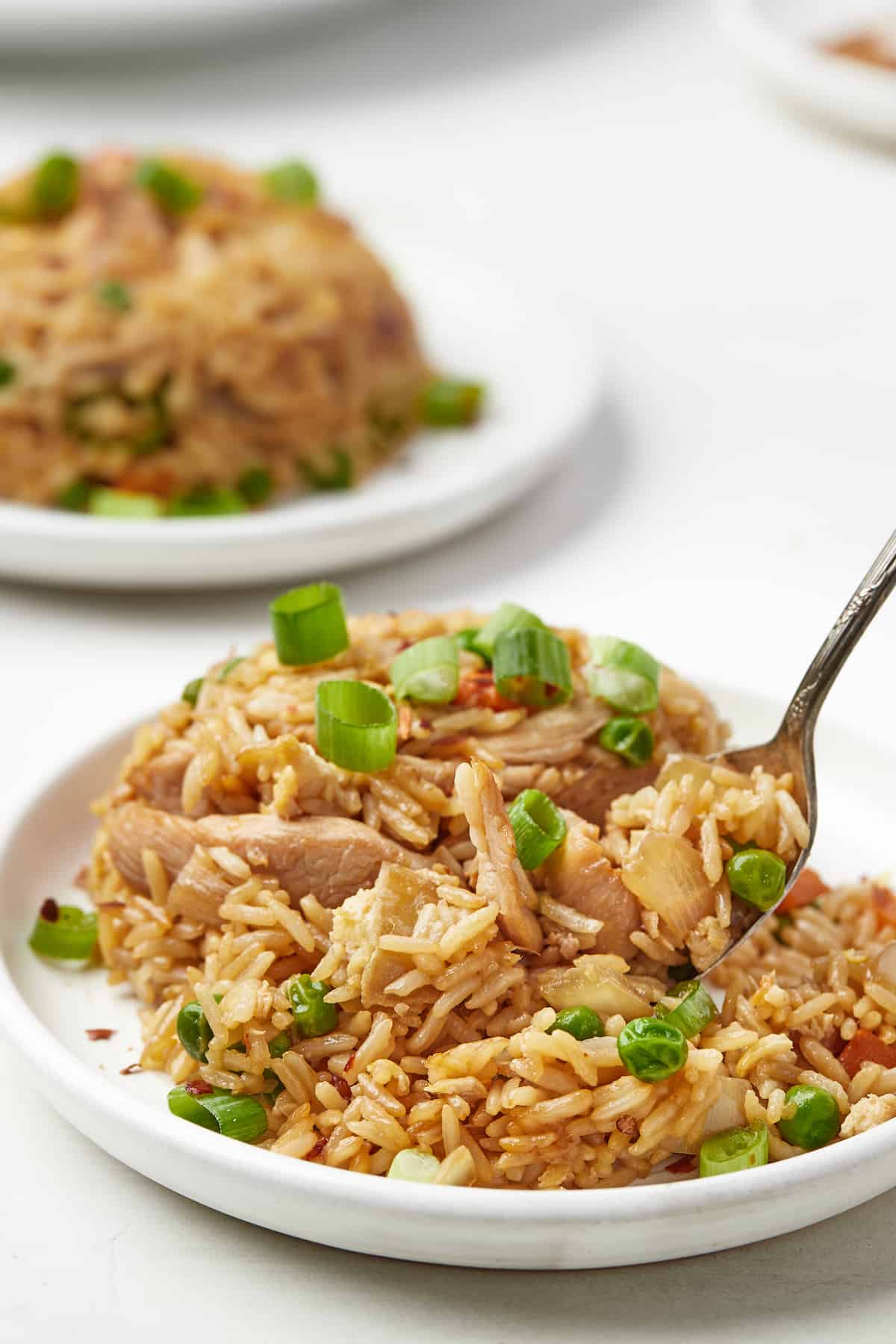 A metal spoon digging into a pile of chicken fried rice on a plate with a second helping on another plate in the background.