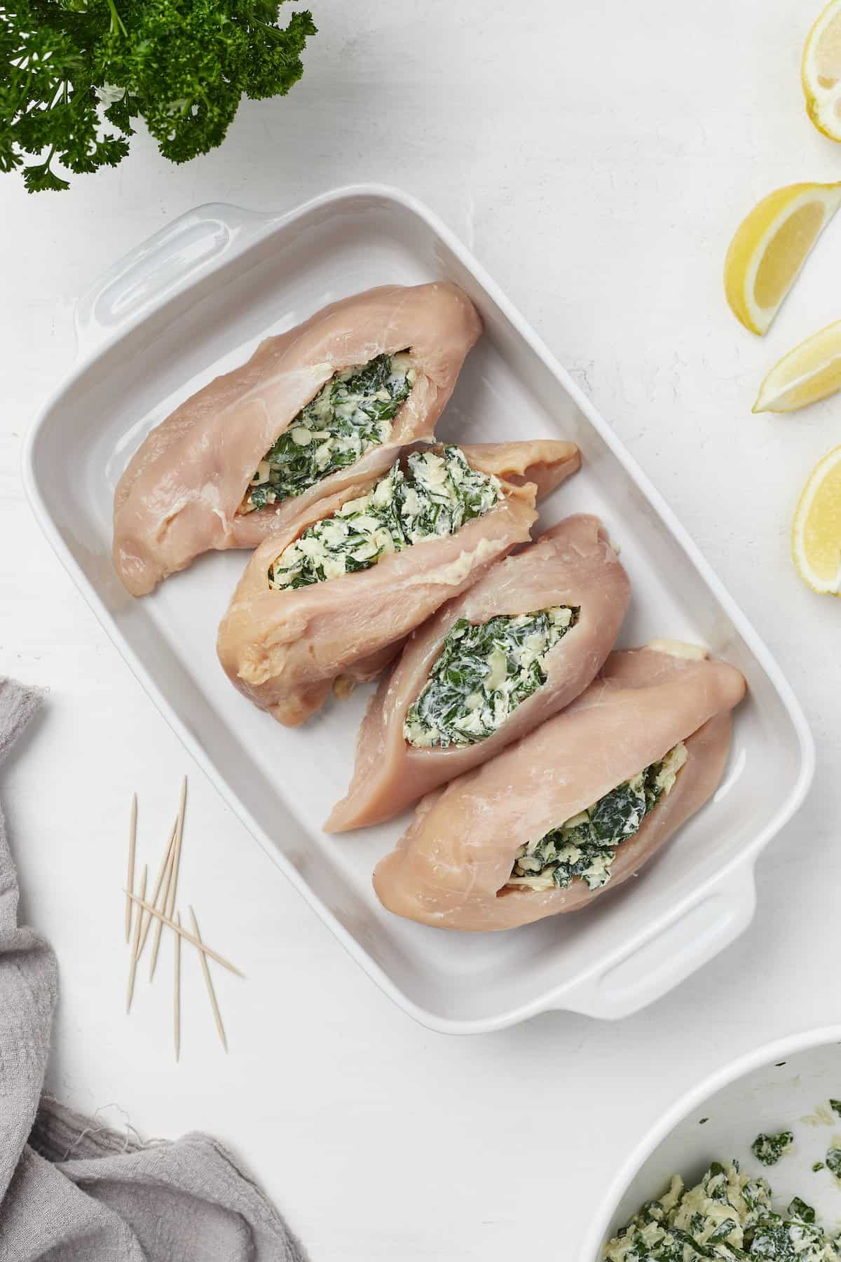 Four stuffed chicken breasts inside of a baking dish with fresh lemon wedges beside it