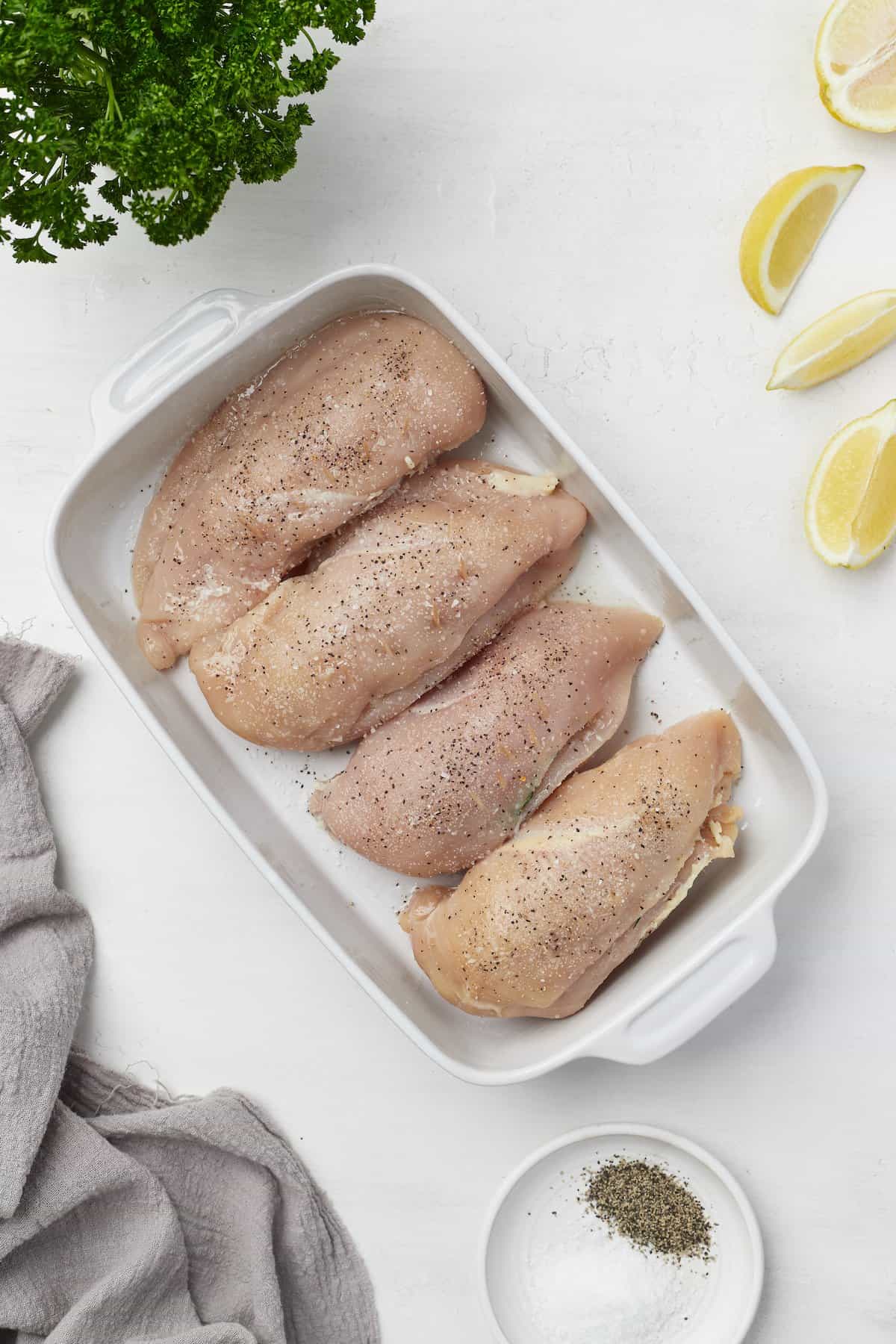 Four stuffed and seasoned chicken breasts in a baking dish with a plate of salt and pepper beside it