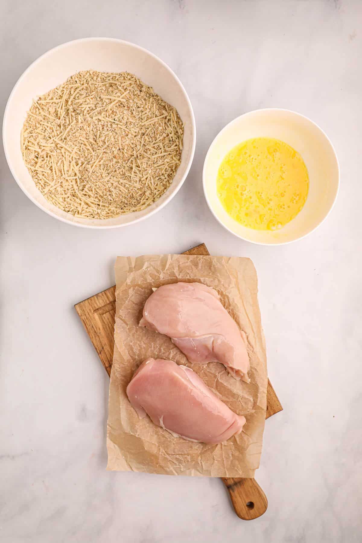 Chicken breasts on a cutting board next to seasoned breadcrumbs and egg wash in bowls.