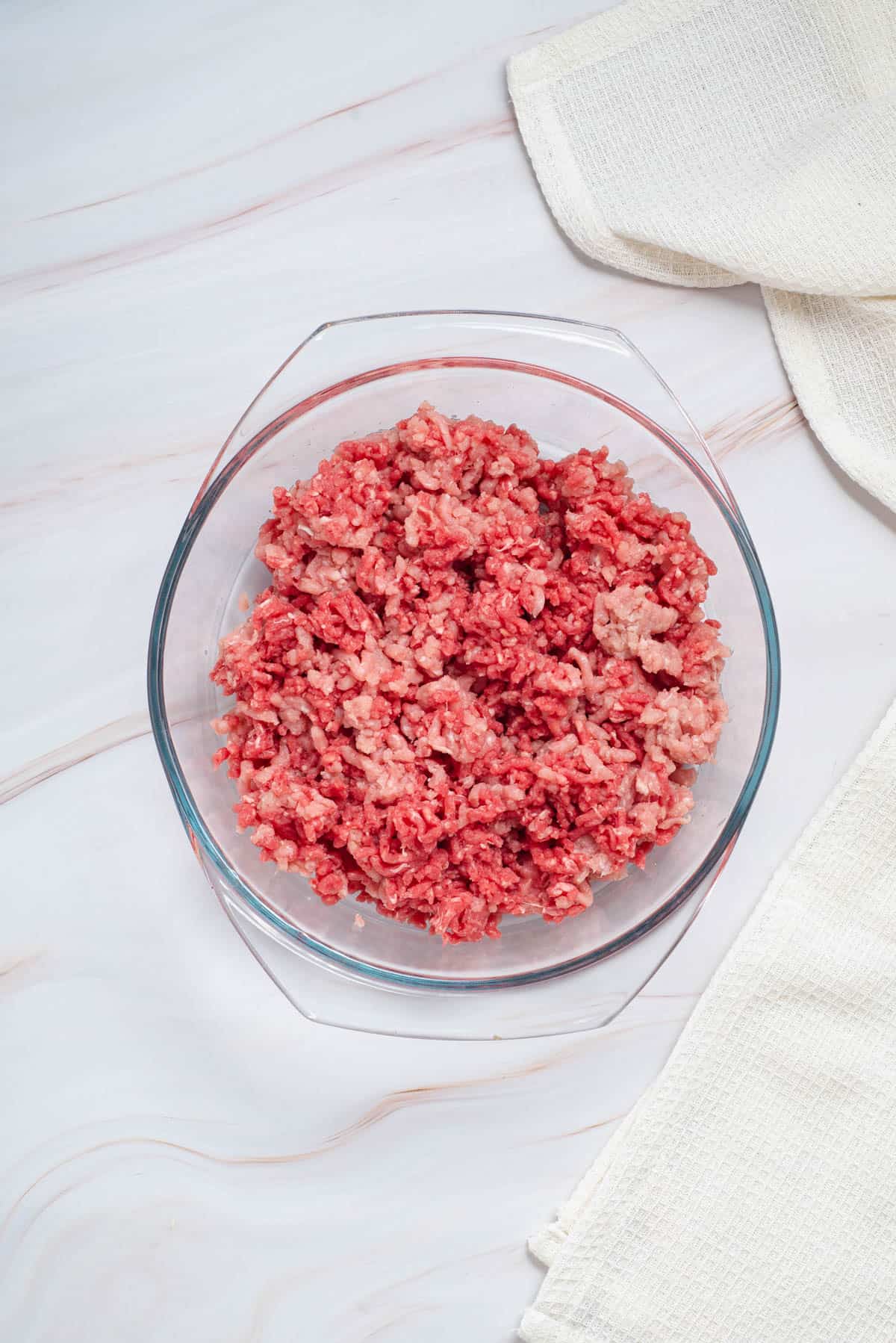 Combining ground pork and ground beef in a large glass bowl for meatballs.