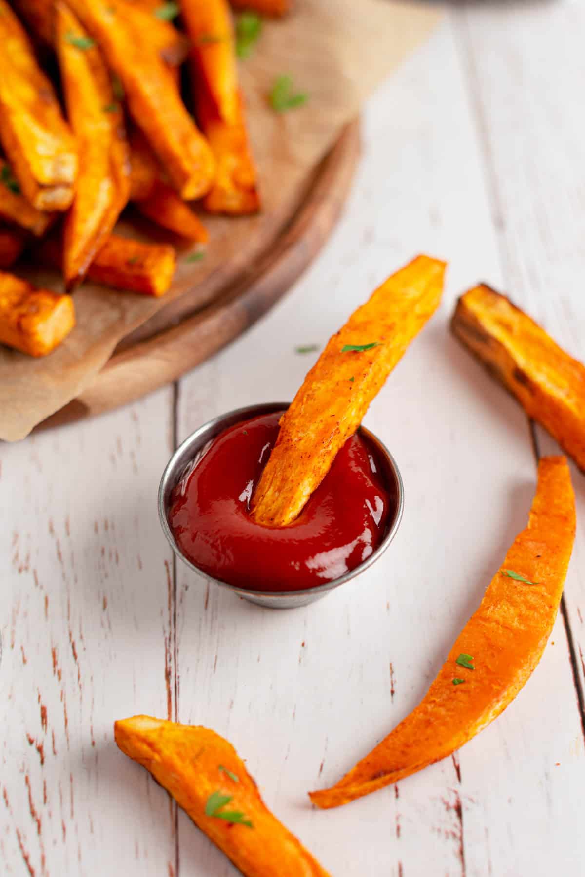 Close up shot of a single sweet potato fry dipped in a serving of ketchup. Other fries are displayed on a plate and near the dip. 