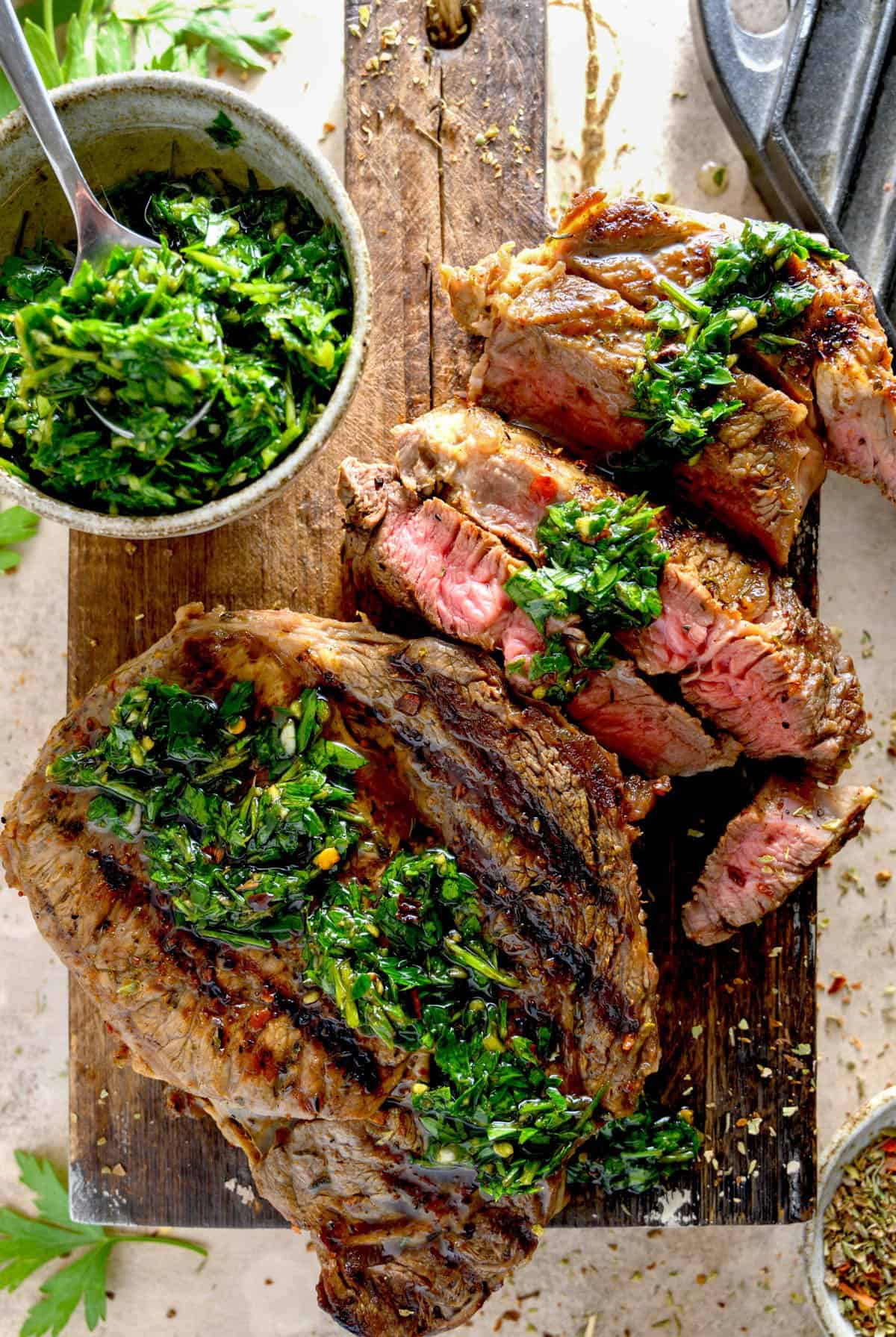 A grilled steak with chimichurri sauce on it and in a bowl next to it on a cutting board.