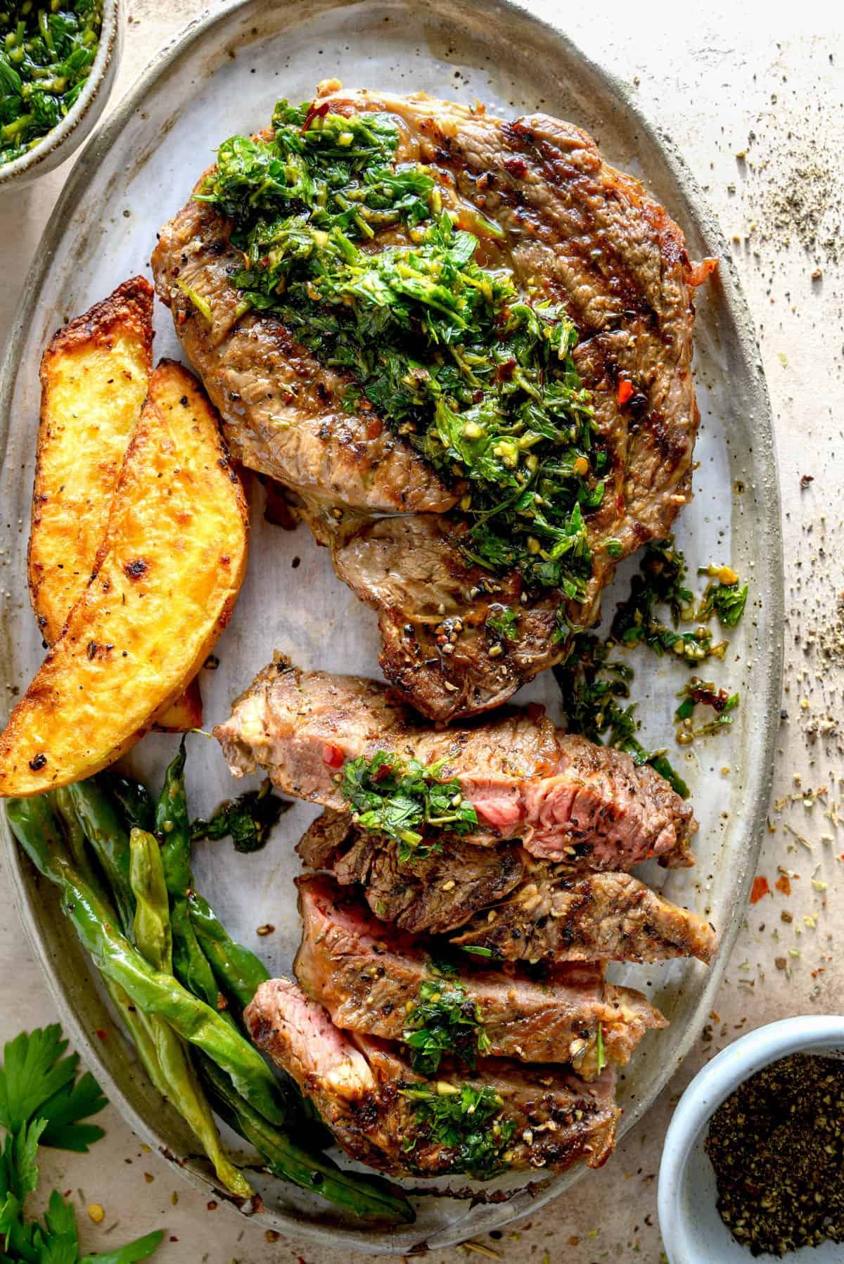 Sliced ribeye steak with chimichurri sauce, vegetables, and potato wedges on a plate.