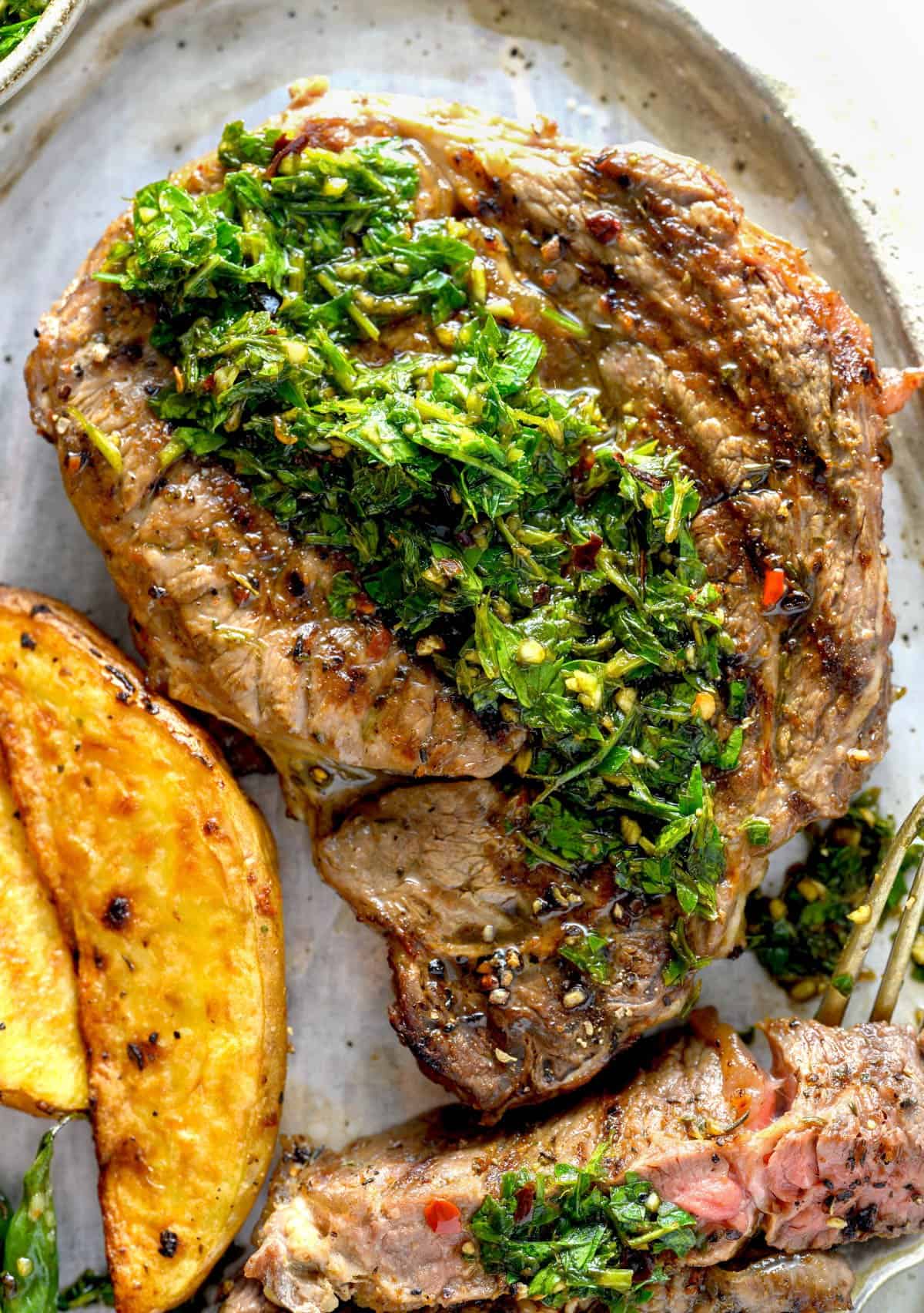 A grilled ribeye steak with chimichurri sauce next to potato wedges.