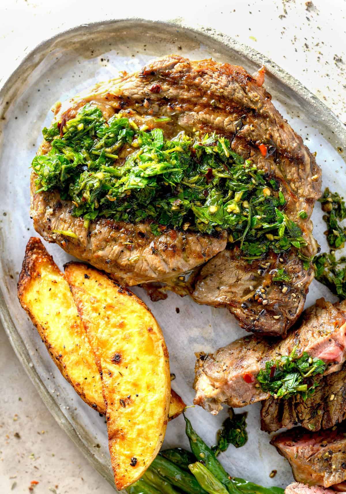 A grilled ribeye steak with chimichurri sauce next to potato wedges.