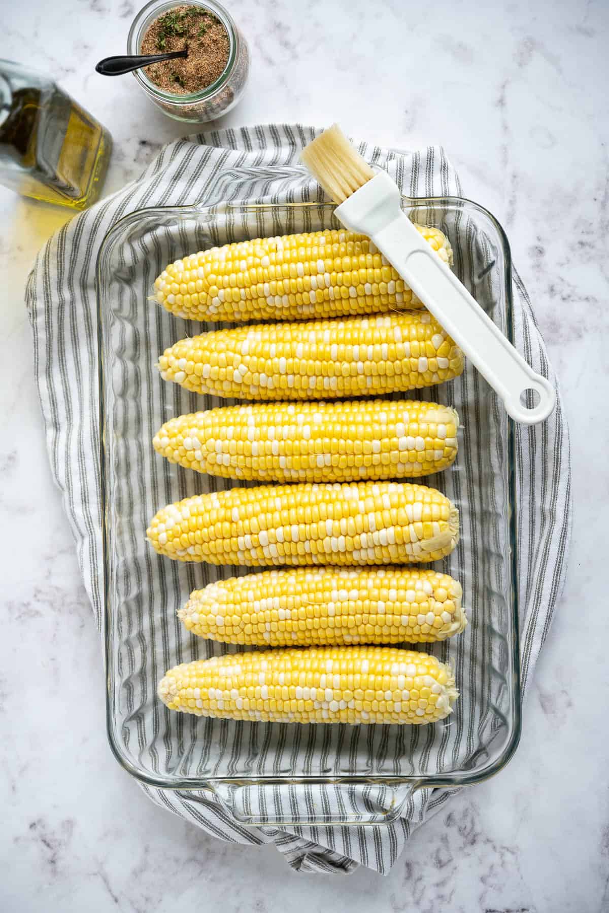 Ears of yellow corn in a glass baking dish with a pastry brush resting on the edge.