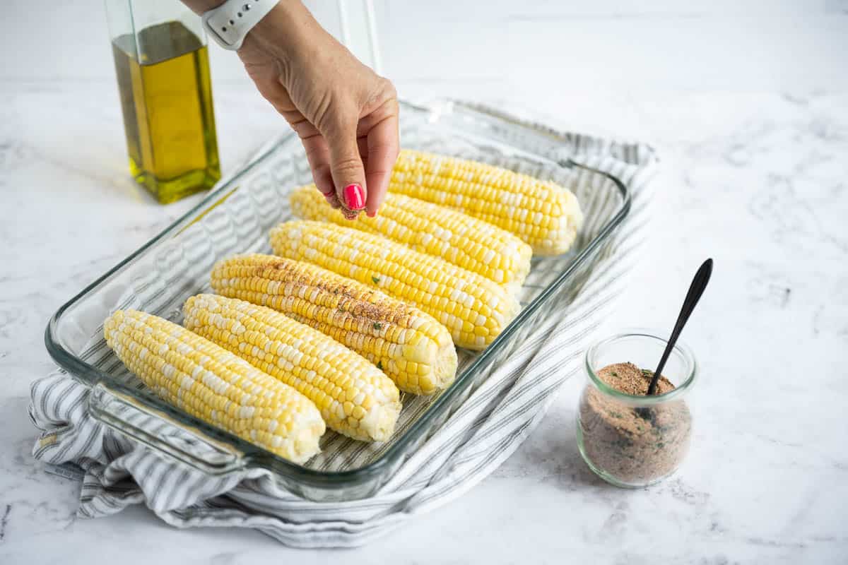 A hand sprinkling seasoning over husked ears of corn in a baking dish.
