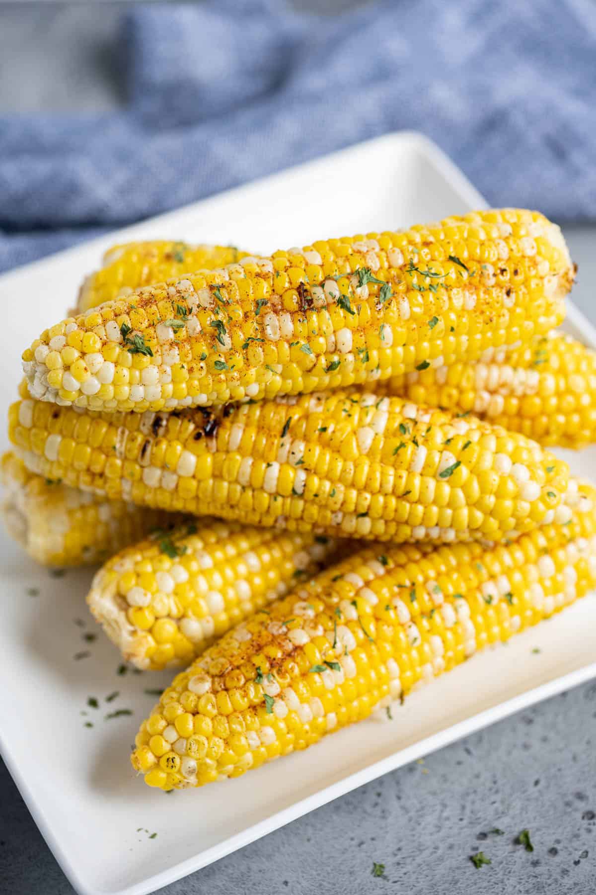 Seasoned and grilled ears of corn on the cob on a plate in front of a blue linen napkin.