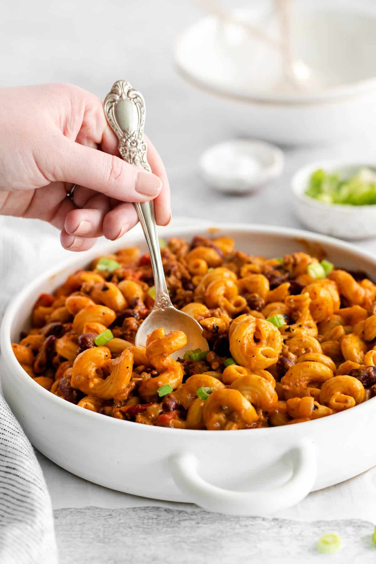 A large white serving dish with chili mac and a hand holding a spoon.