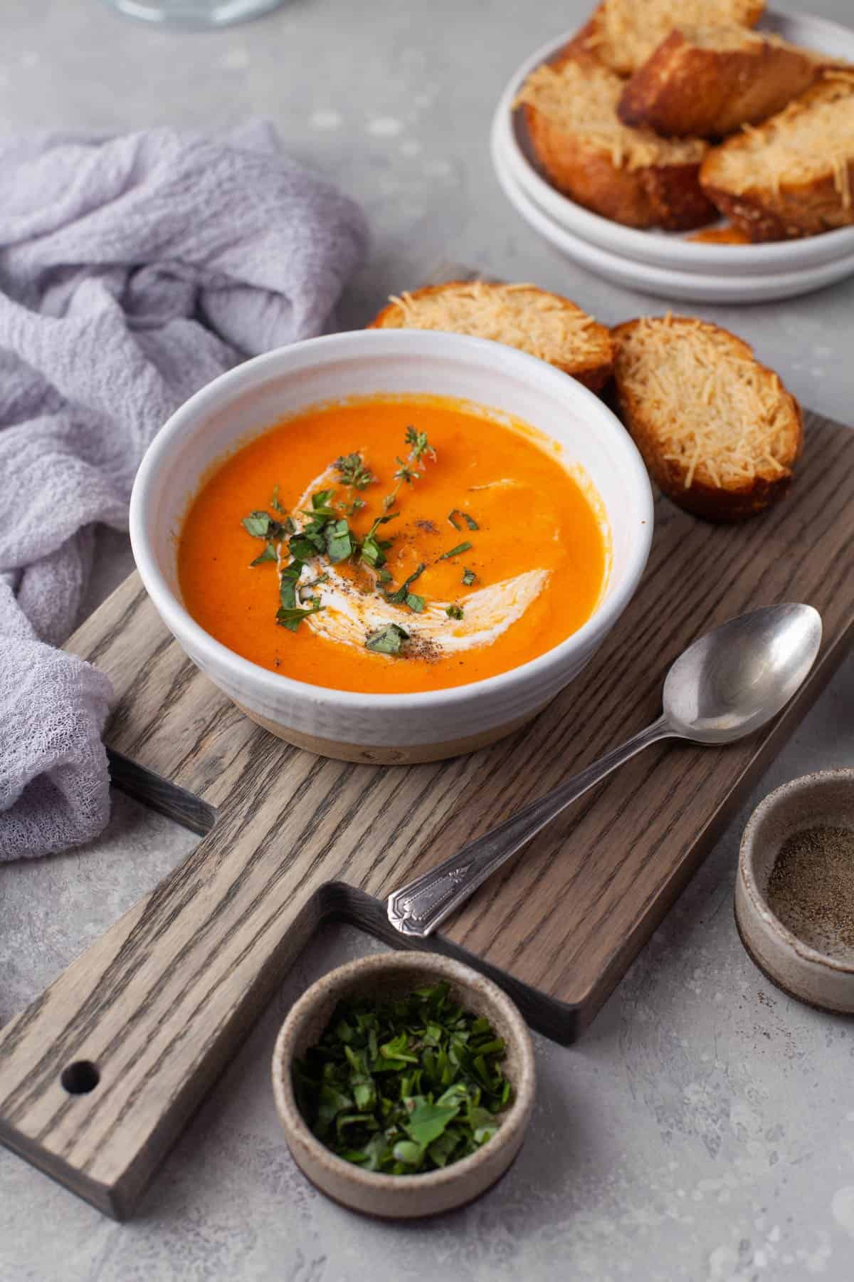 A bowl of creamy tomato soup on a wooden cutting board next to a bowl of fresh herbs, a linen napkin, and toasted bread.
