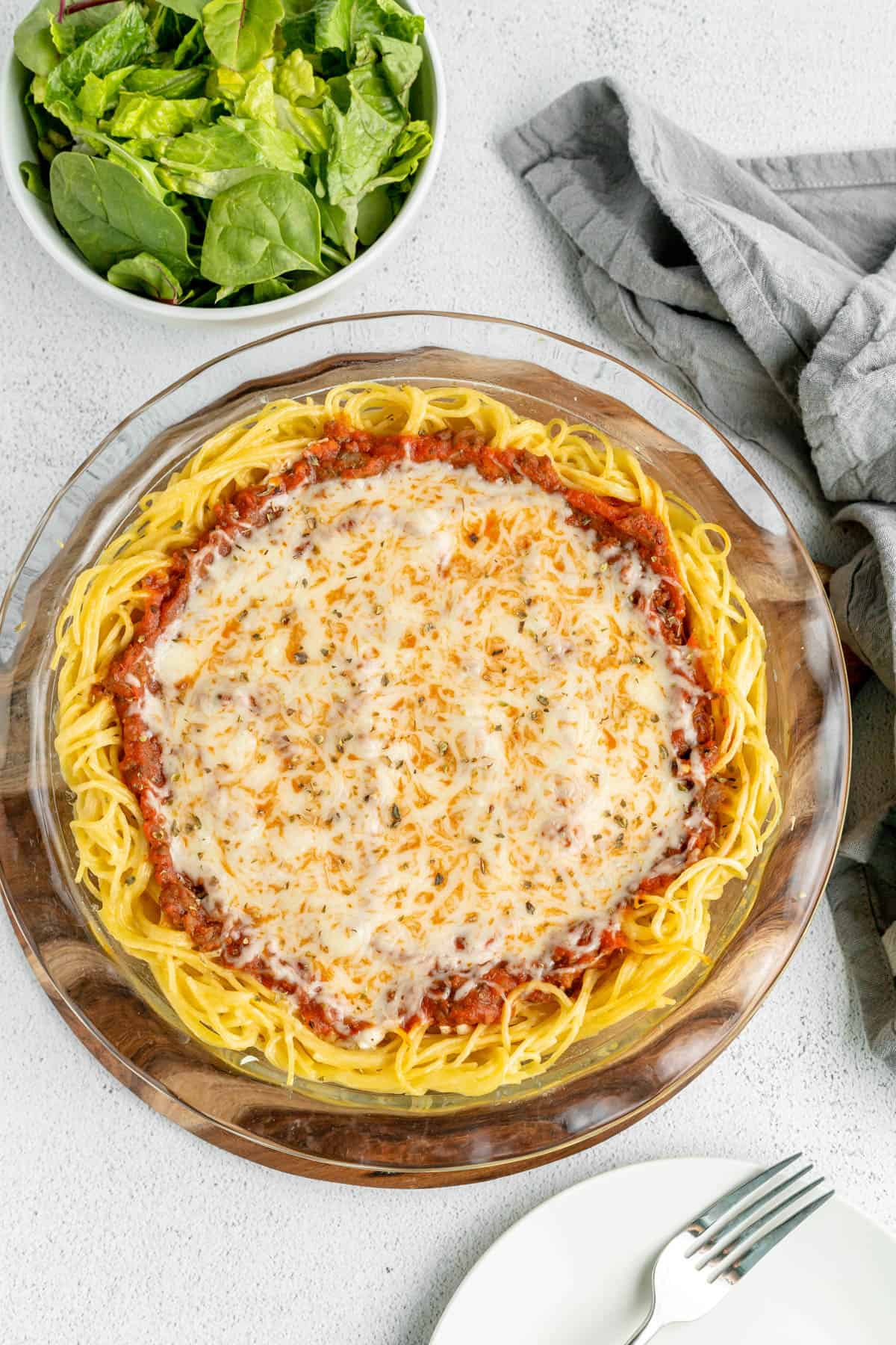 A cooked spaghetti pie next to a bowl of lettuce.