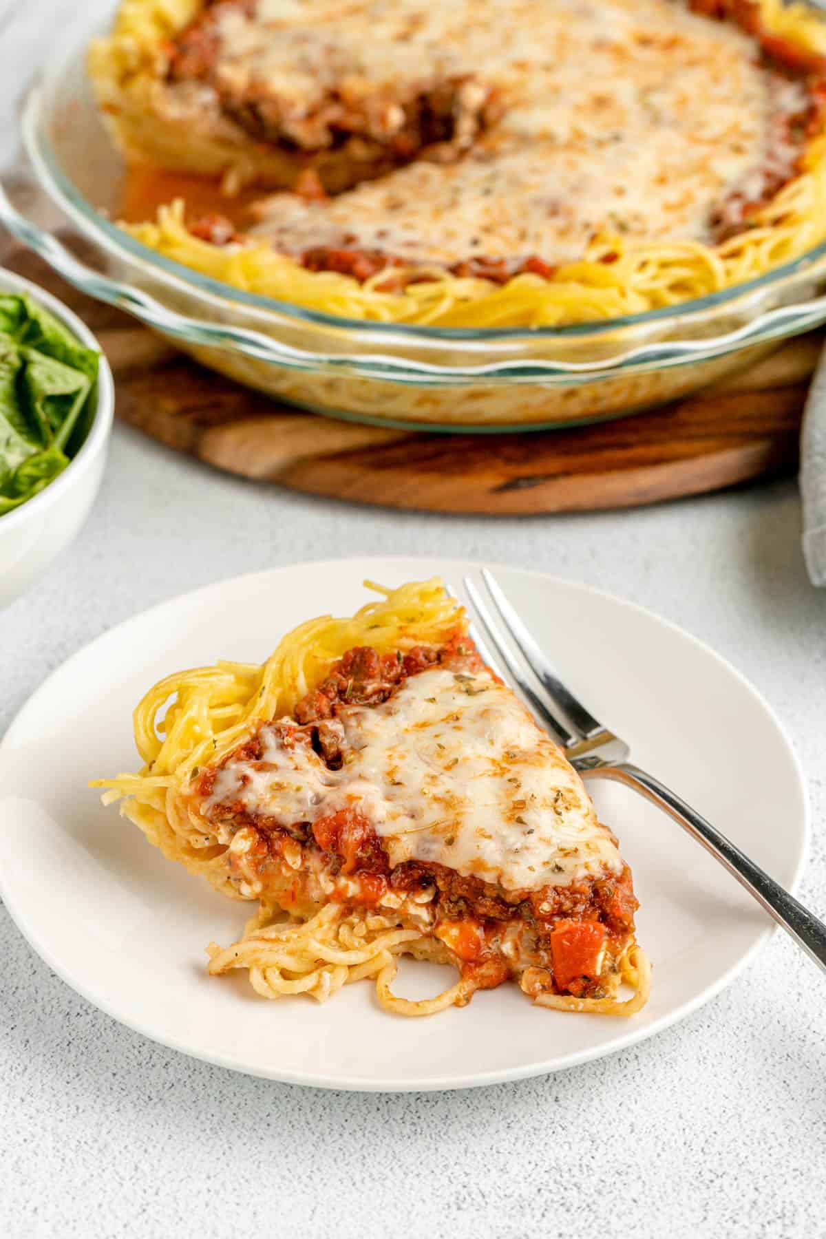 A slice of spaghetti pie on a white plate with a fork in front of the pie dish it was baked in.