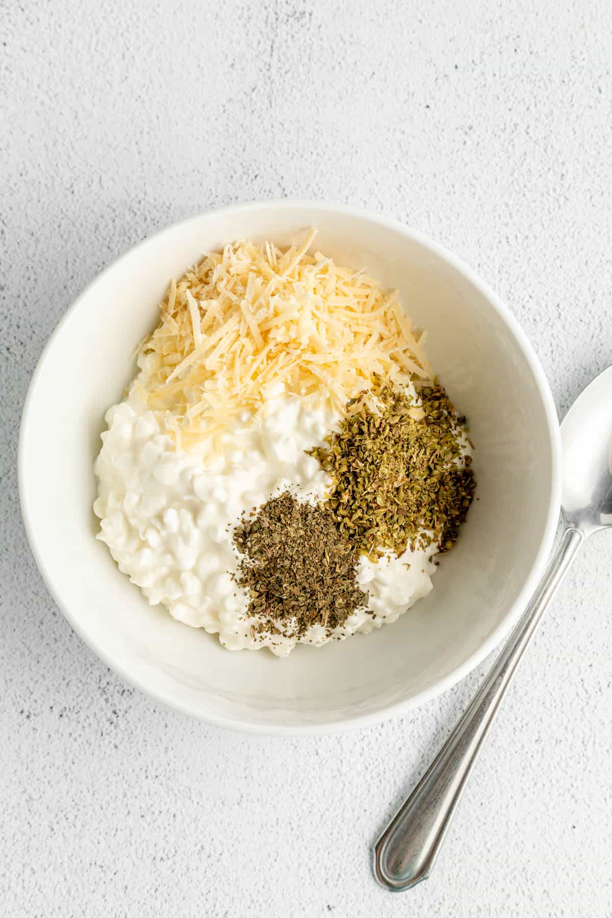 Cottage cheese, grated parmesan cheese, and seasonings in a white bowl next to a spoon.