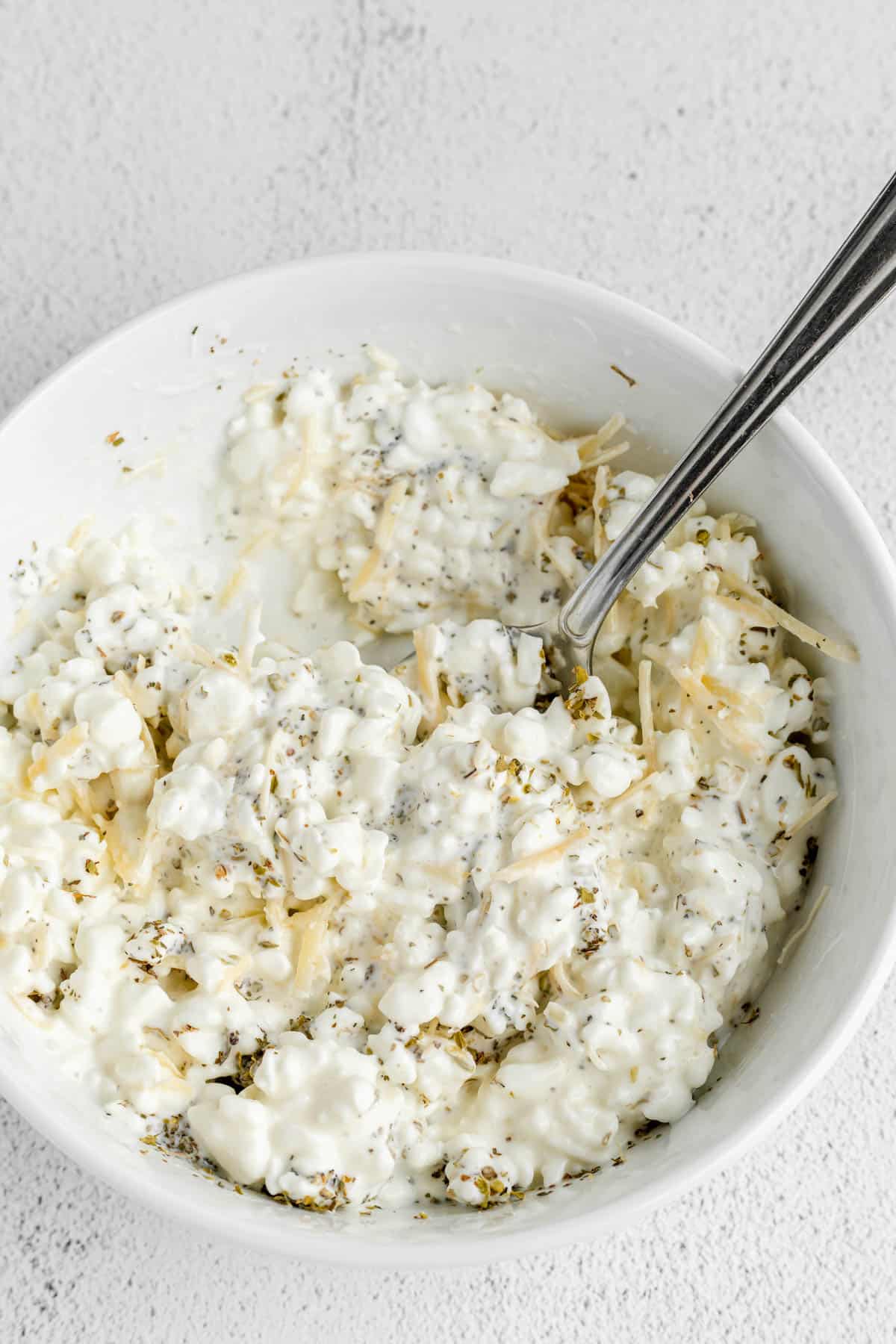 Mixed cottage cheese and spice mixture for making spaghetti pie in a white bowl with a spoon.