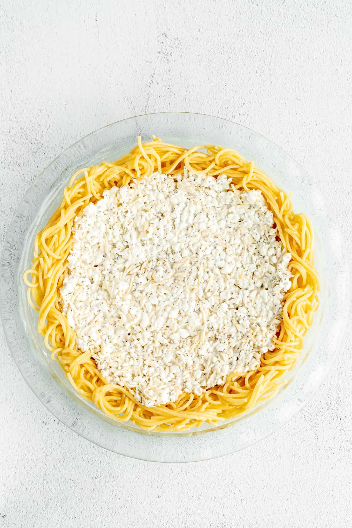 Adding cottage cheese mixture to cooked spaghetti noodles in a glass pie dish.
