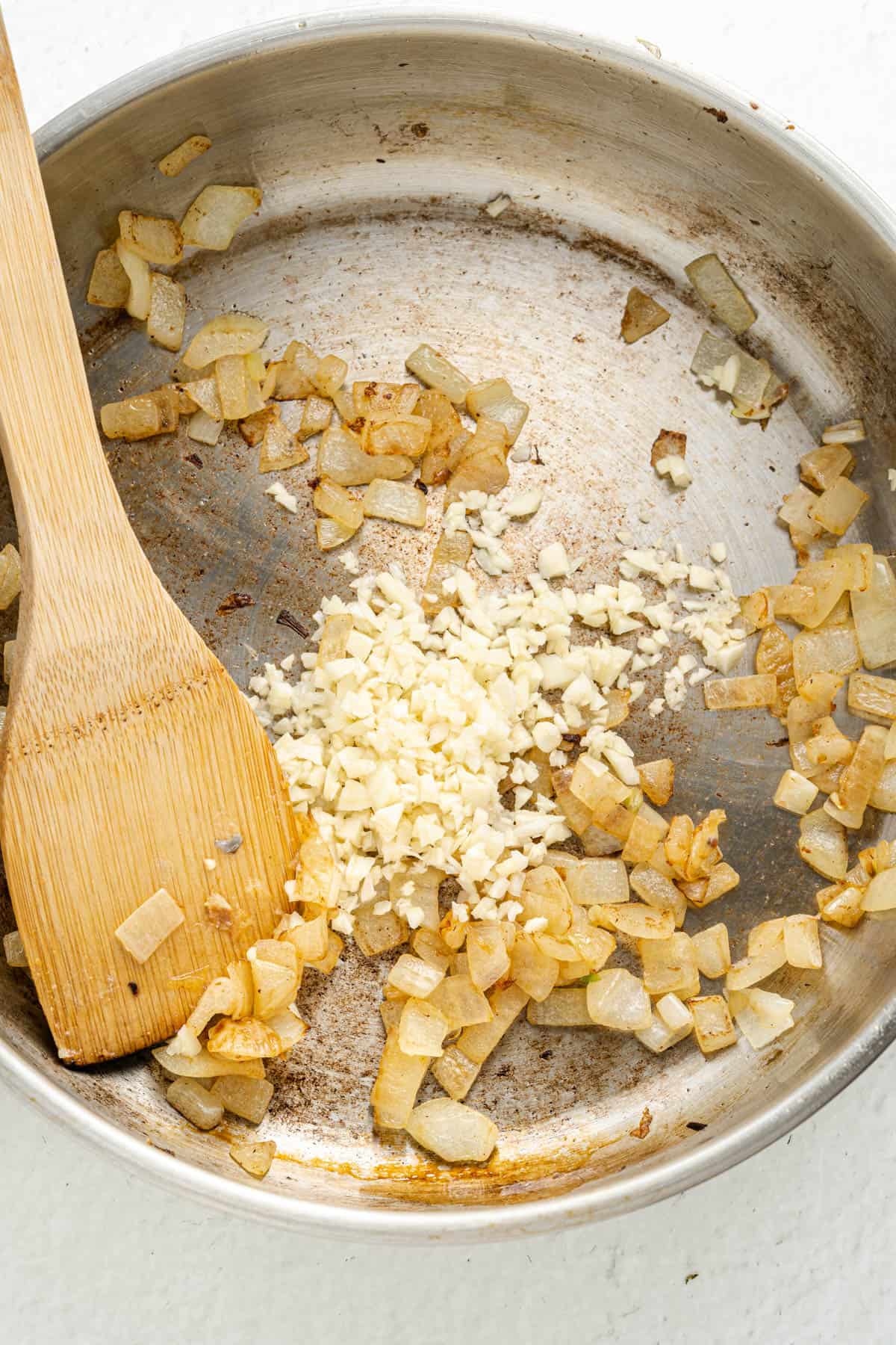 Adding minced garlic to a pan of sauteed onions with a wooden spoon.