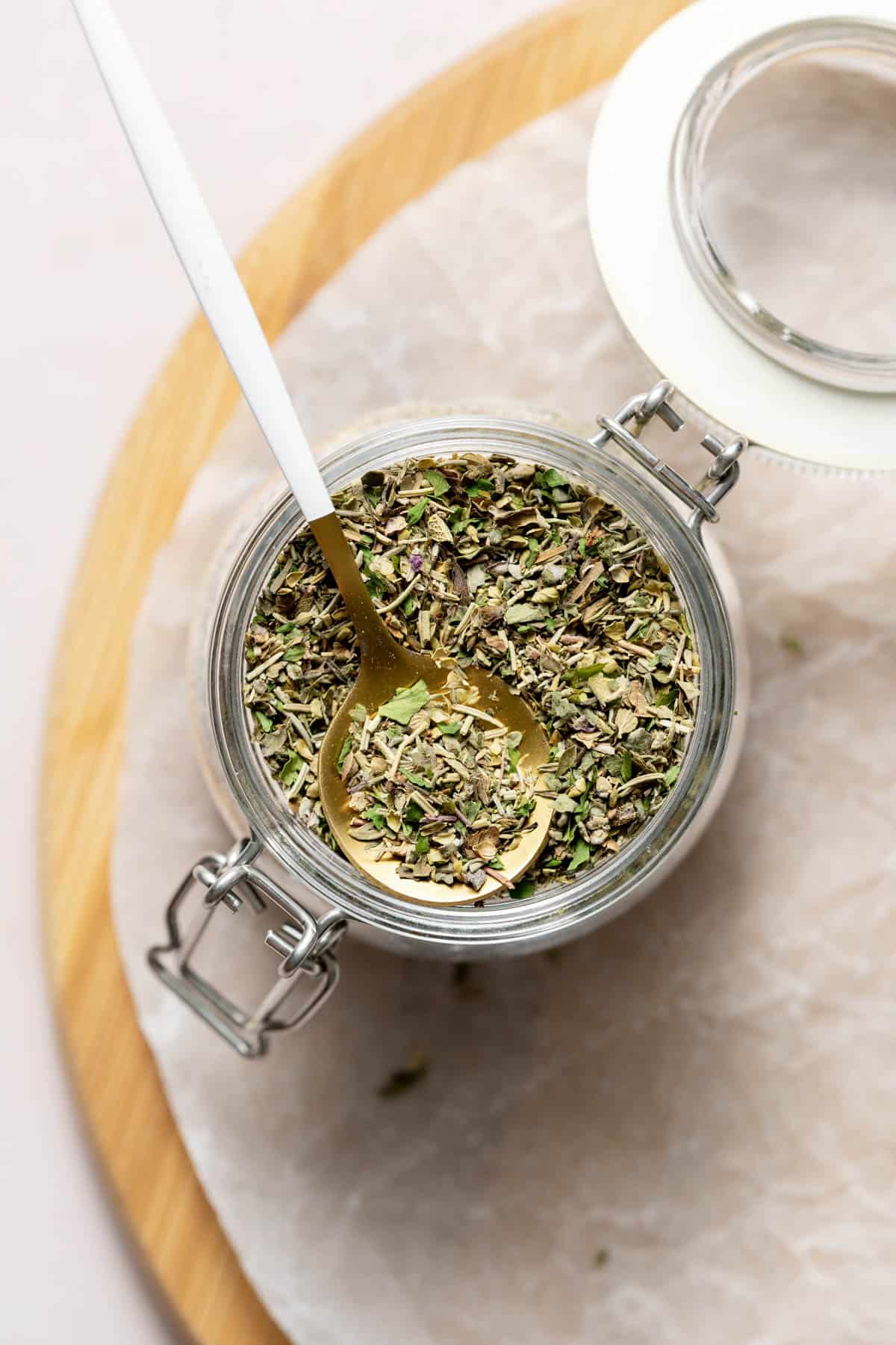 A gold and white spoon with homemade Italian seasoning in a jar of the spice blend.