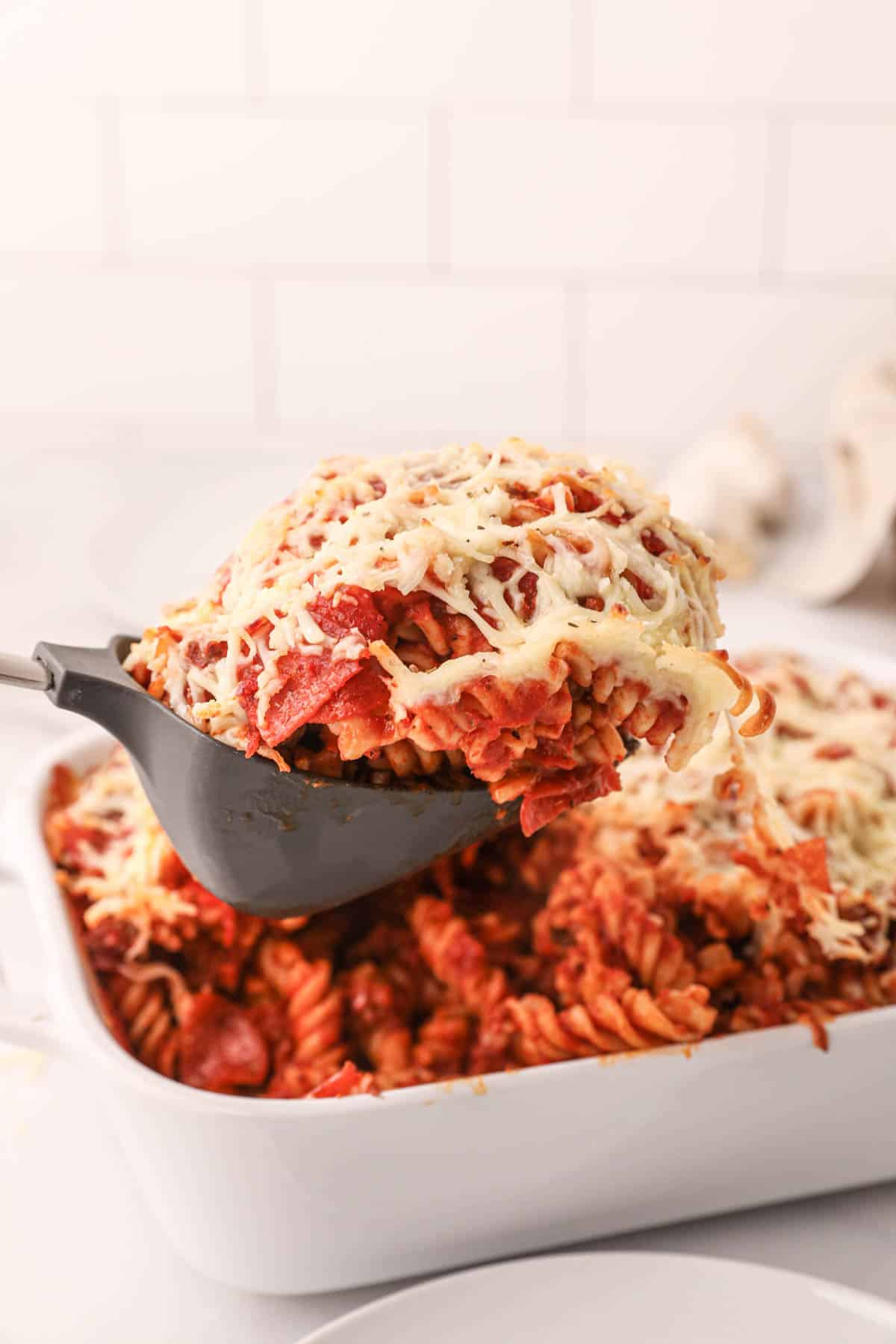 A large spoon lifting a scoop of pizza pasta from a casserole dish.