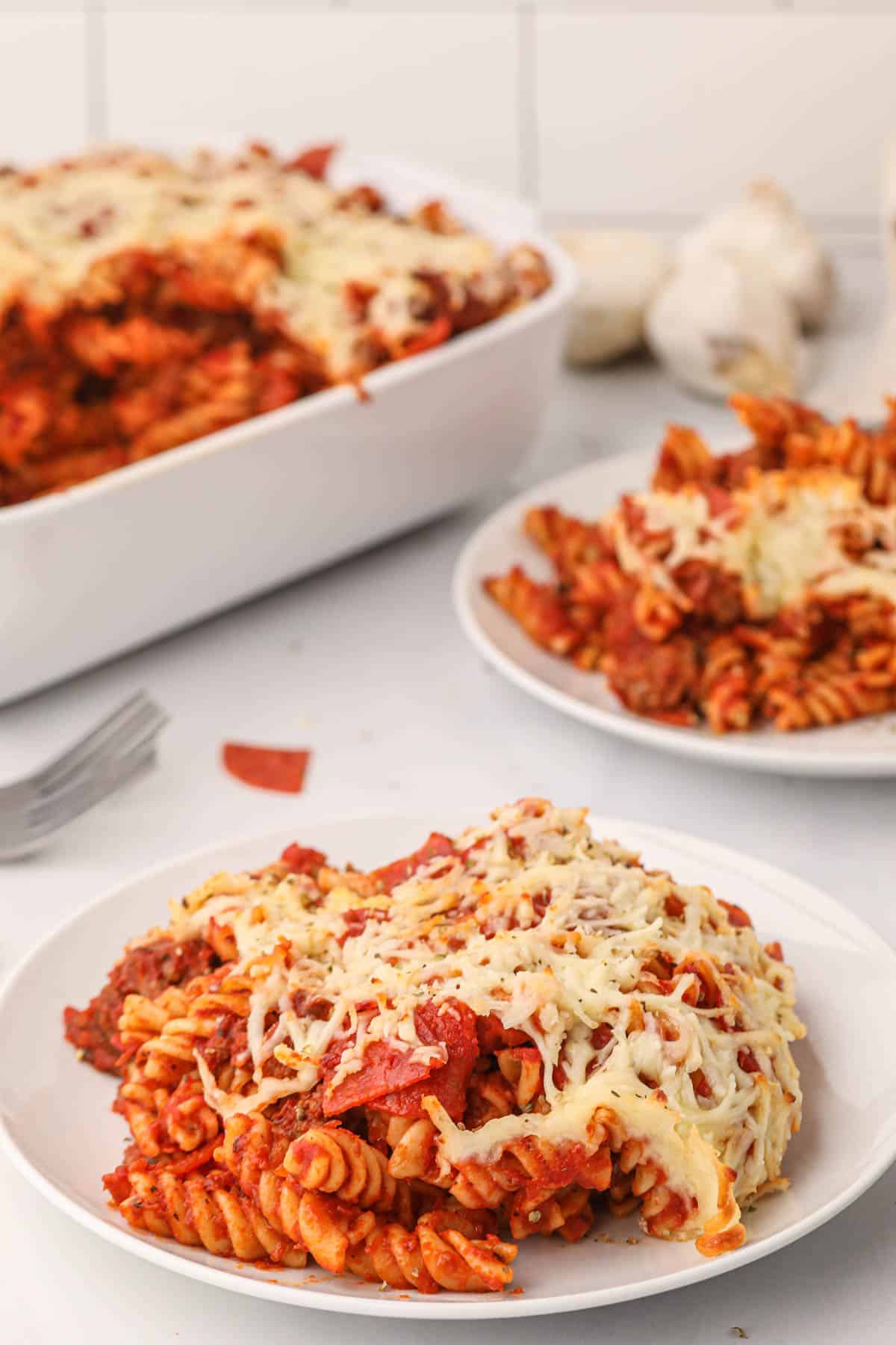 A serving of pizza pasta on a white plate in front of the rest of the casserole and another serving.