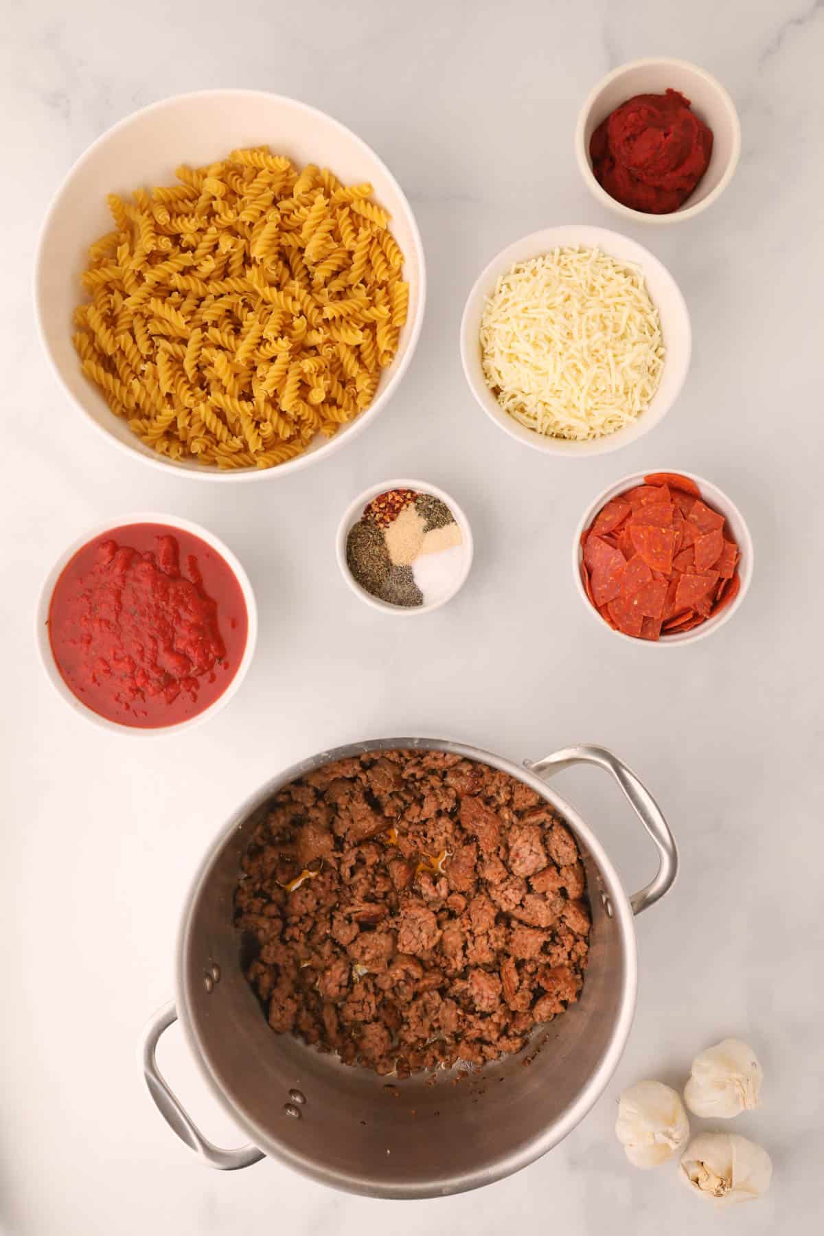 Cooked Italian sausage in a large pot next to the remaining ingredients for making pizza pasta.