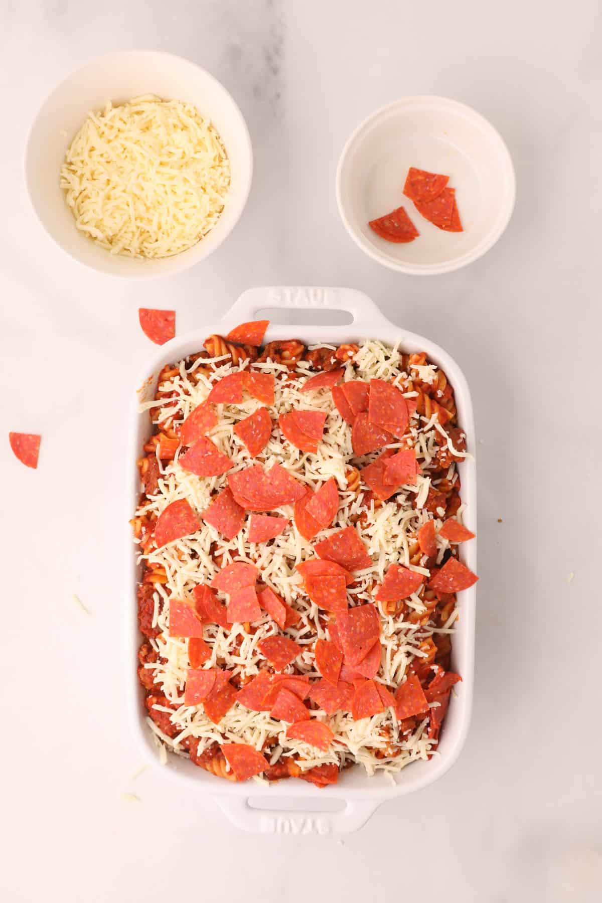 Adding layers of cheese and pepperoni to pizza pasta casserole in a white baking dish.