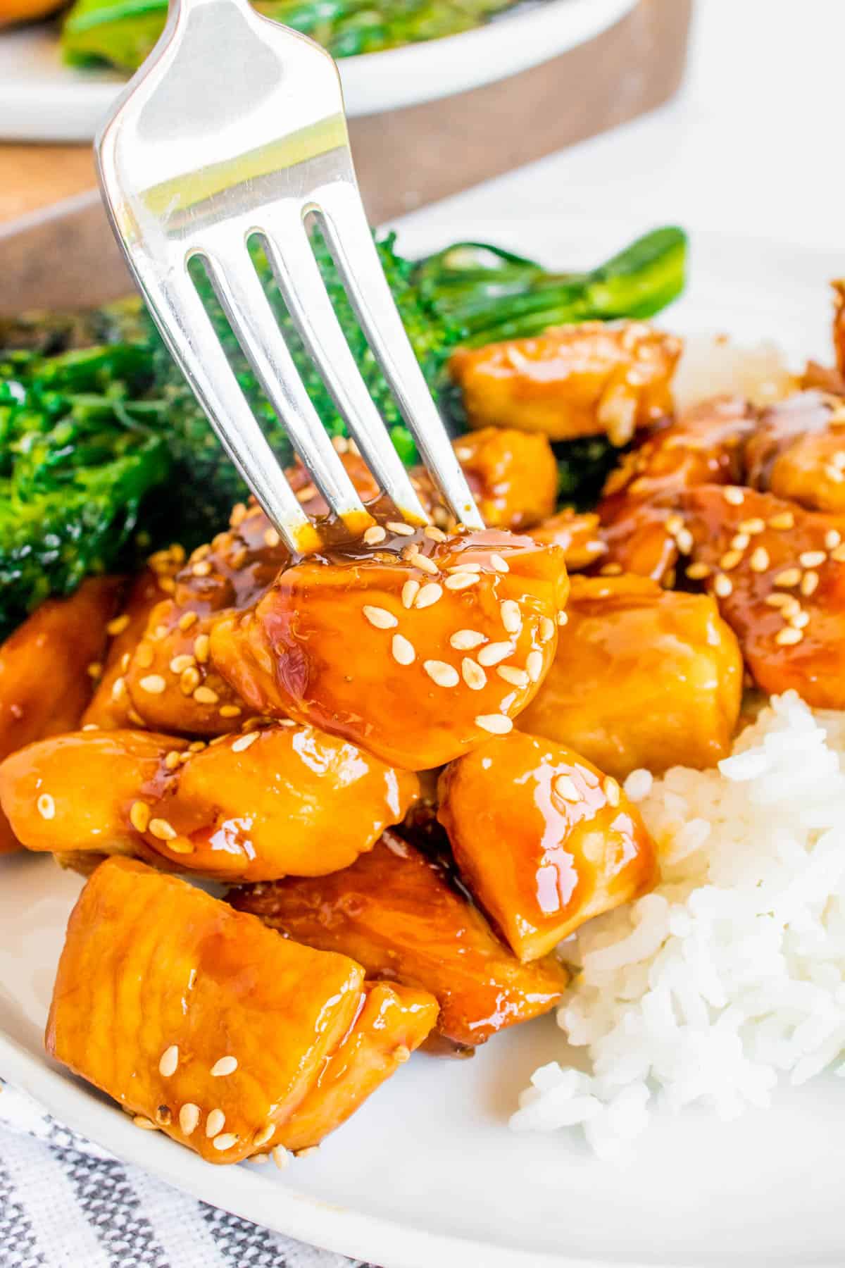 Close up of Teriyaki Chicken served on a plate with white rice and broccolini in background. A fork is visible, picking up a piece of chicken. 