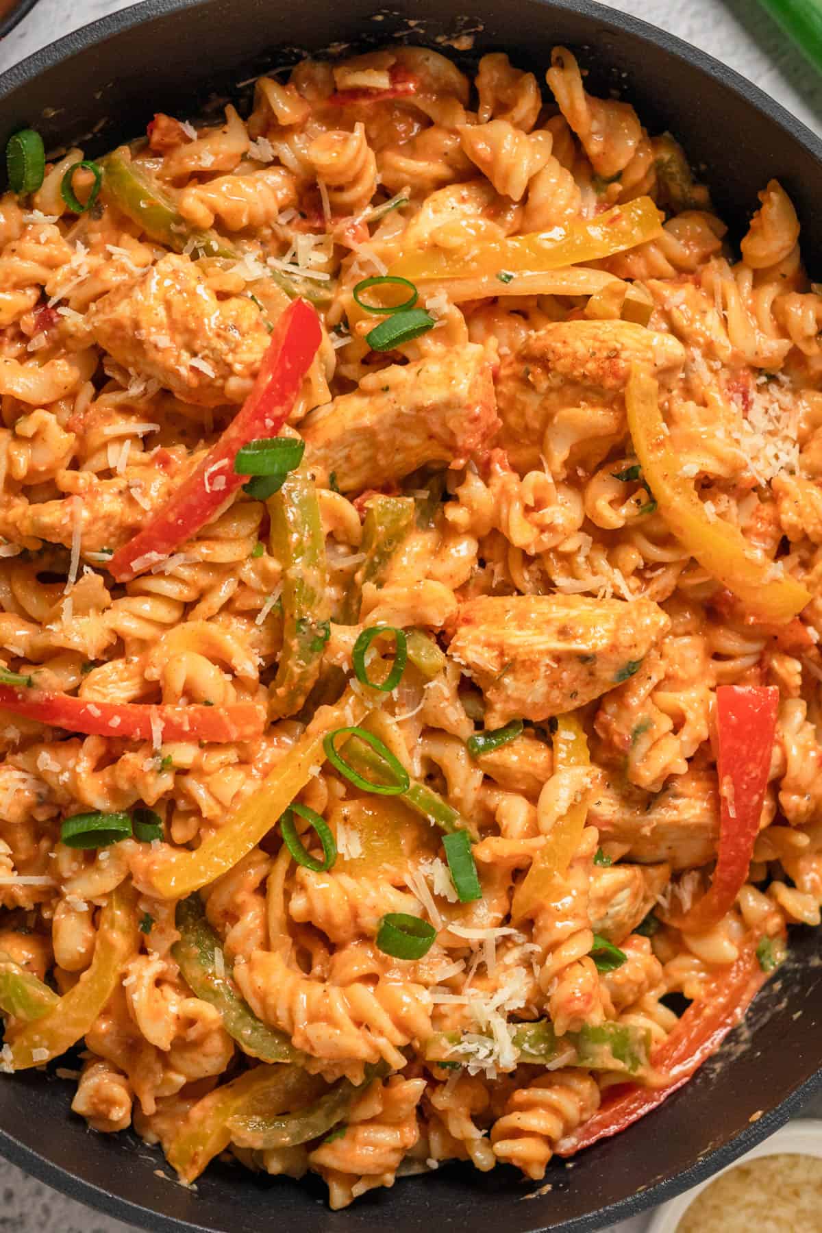 A close image of fusilli pasta with cooked chicken pieces and sliced red, yellow, and green bell peppers.